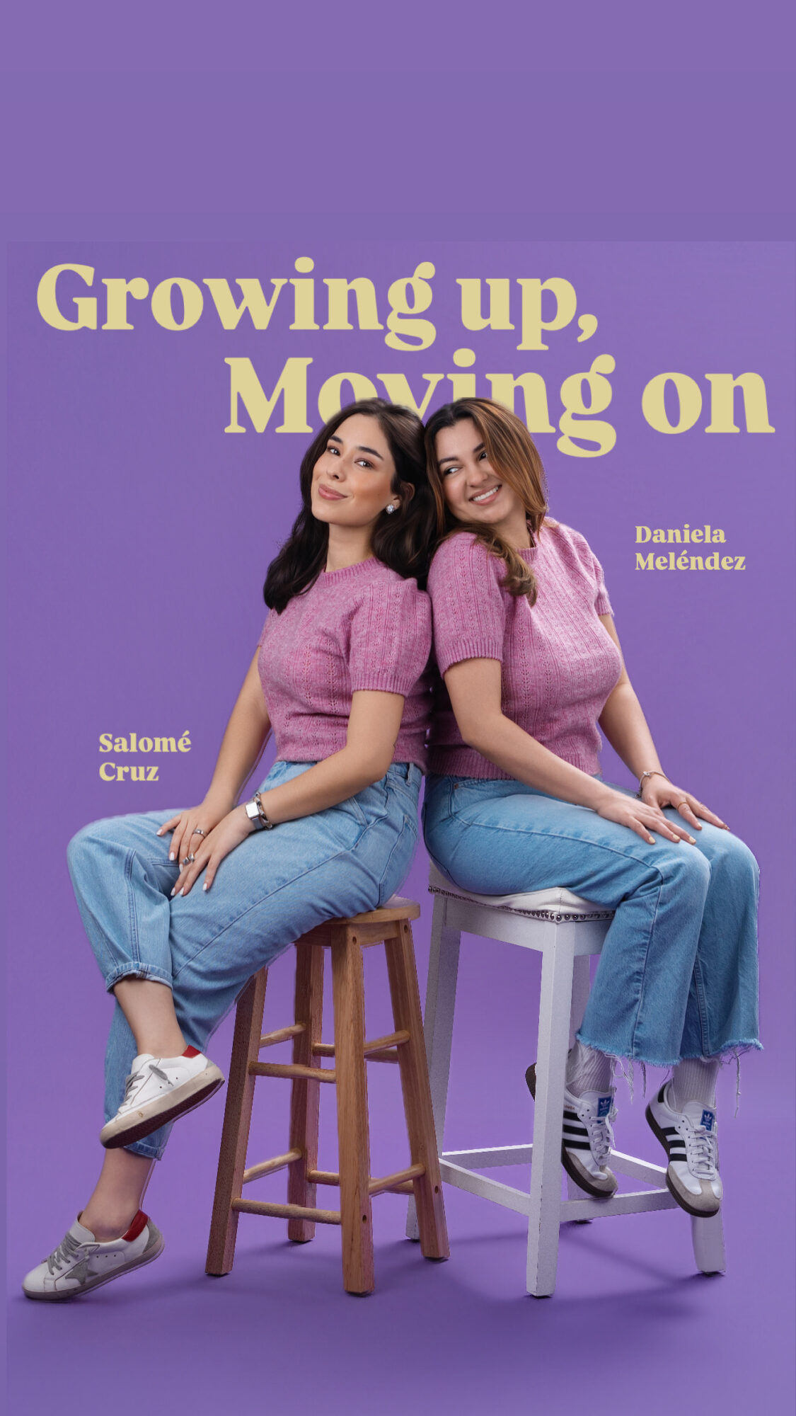 Two women sit on stools, smiling, wearing purple tops and jeans, against a purple background.