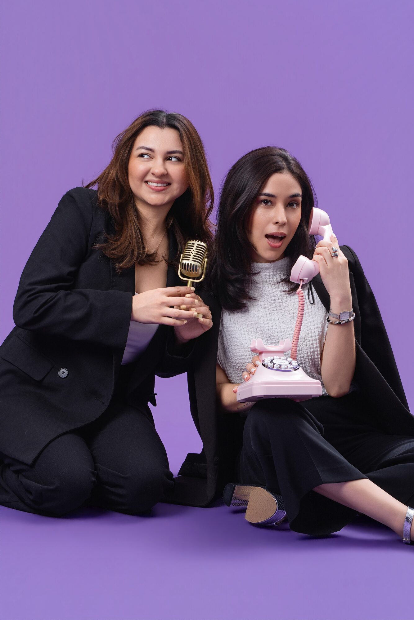 Two women sitting on the floor with a purple background, one holding a vintage microphone and the other a rotary phone, smiling and talking.