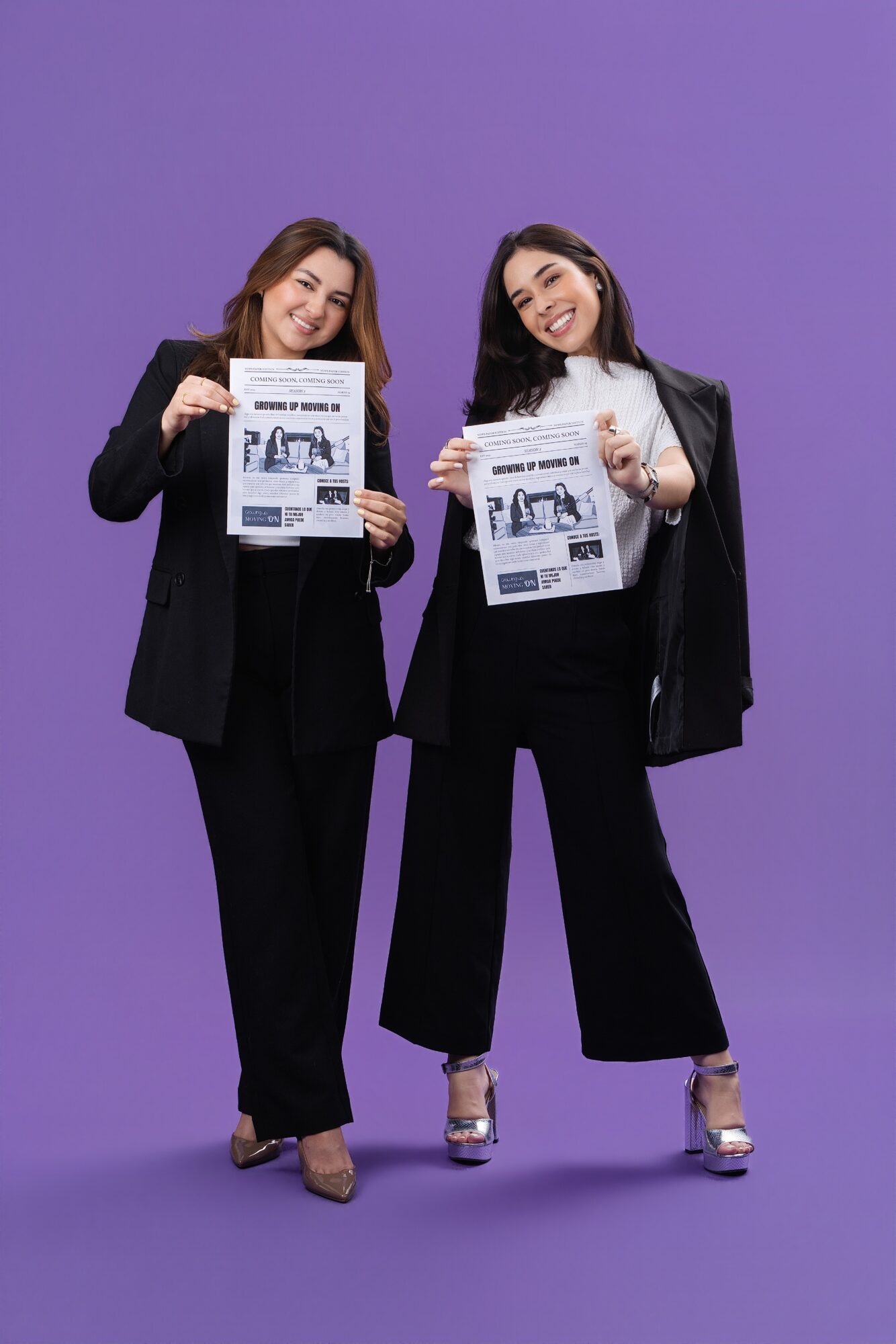 Two women in black suits holding newspapers, smiling, standing against a purple background.