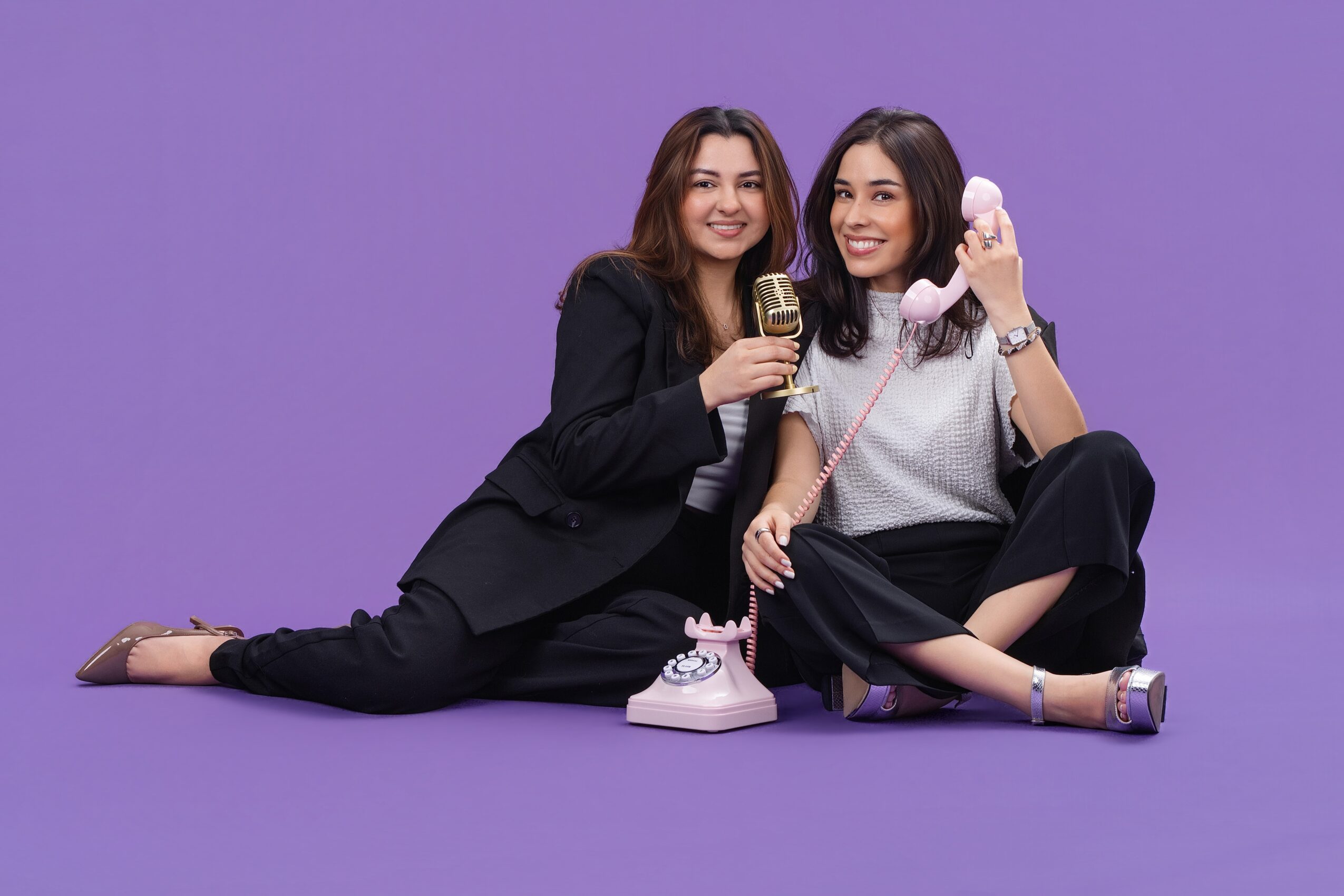 Two women sit on the floor with a purple background, one holding a microphone and the other a phone, smiling.