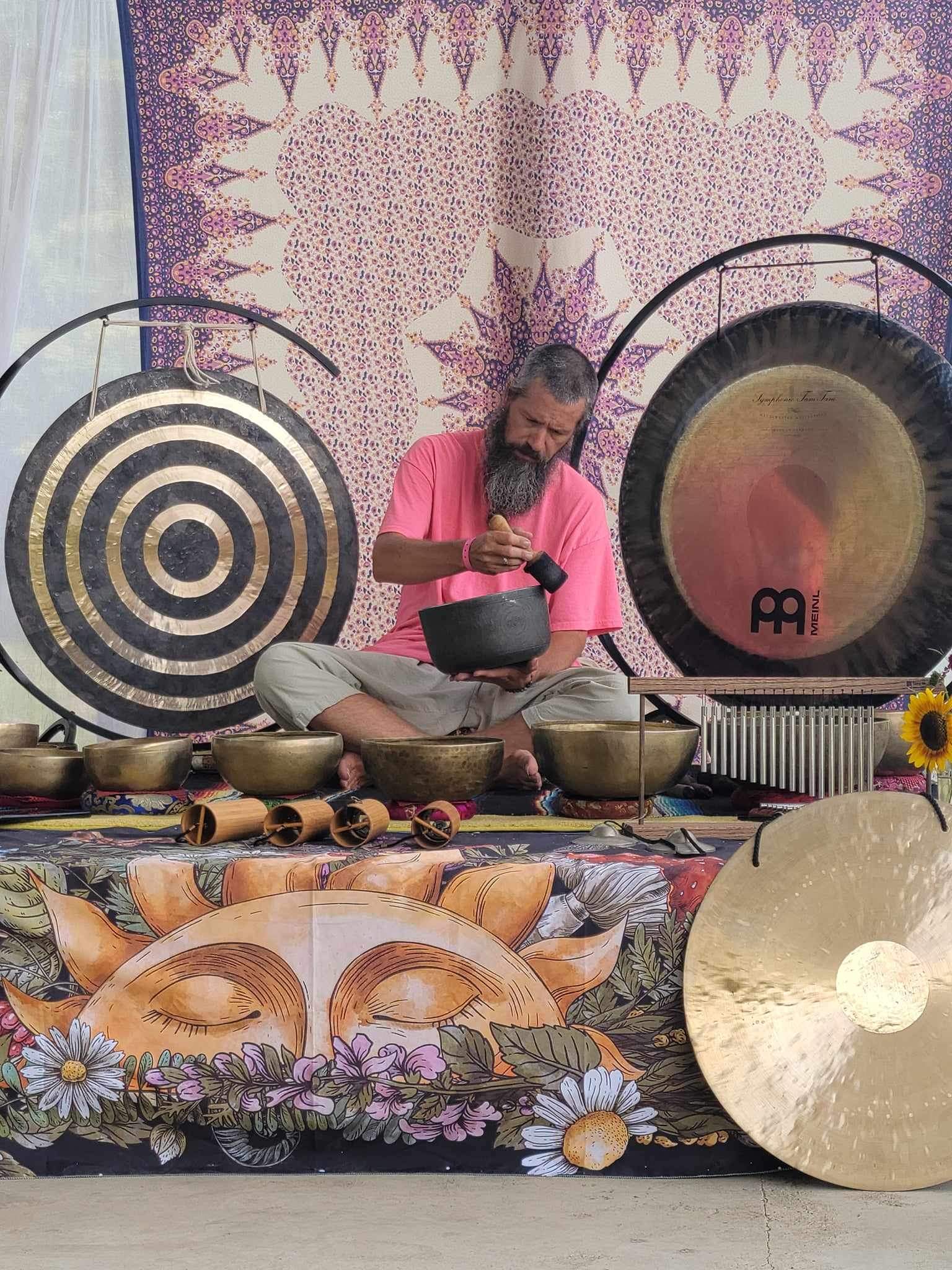 Man in pink shirt playing a gong surrounded by bowls and decorative items, with a colorful backdrop and floral decorations.