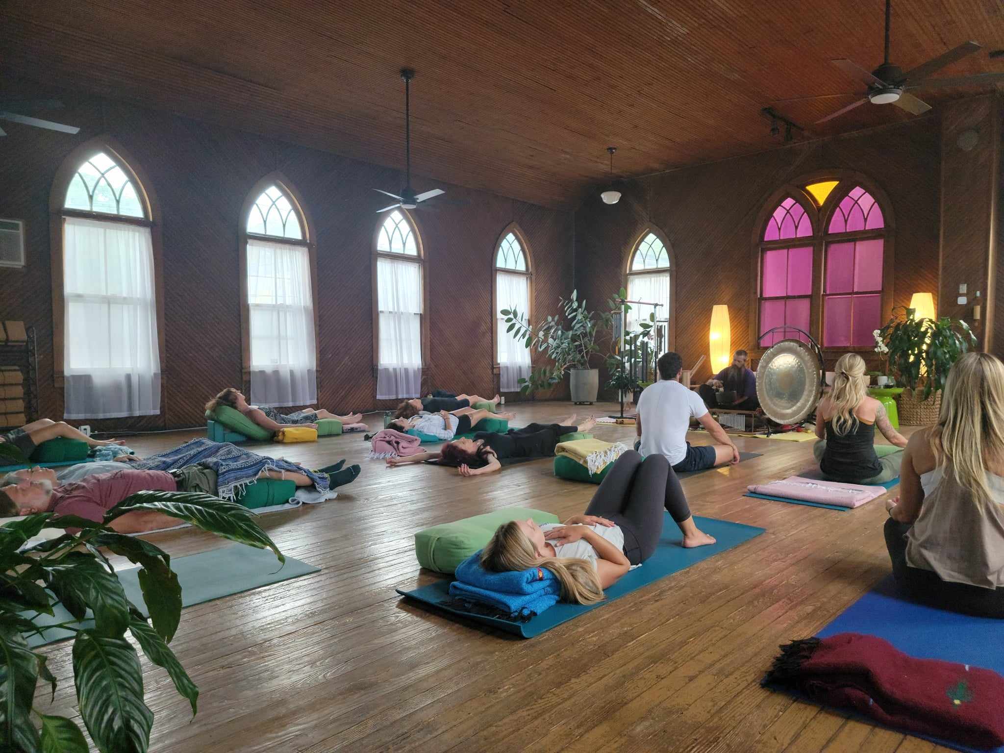 People practicing yoga or meditation on mats in a spacious room with arched windows and wooden floors.