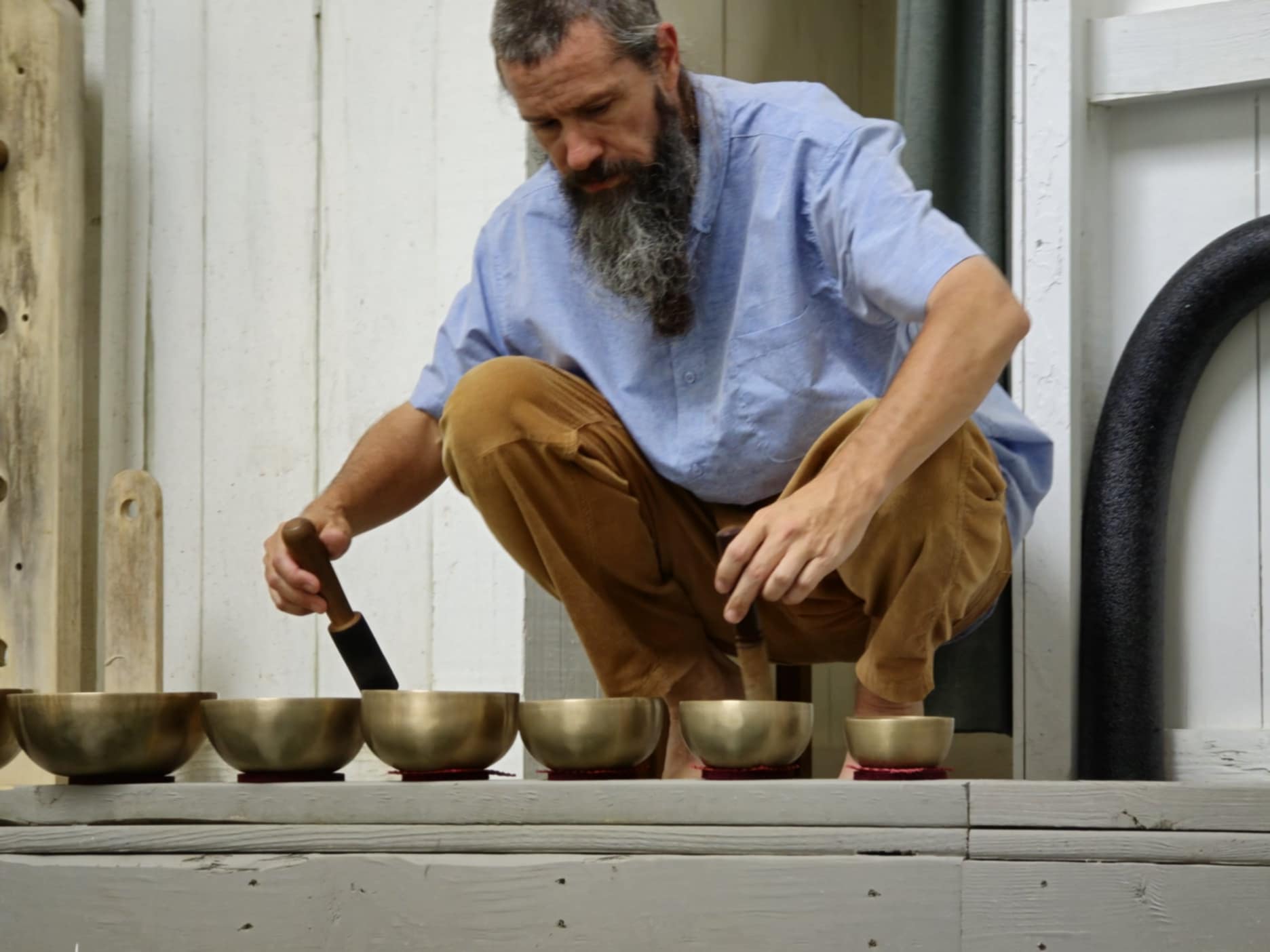 Man with a beard crouching outdoors, arranging bowls on a white surface, wearing a blue shirt and brown pants.