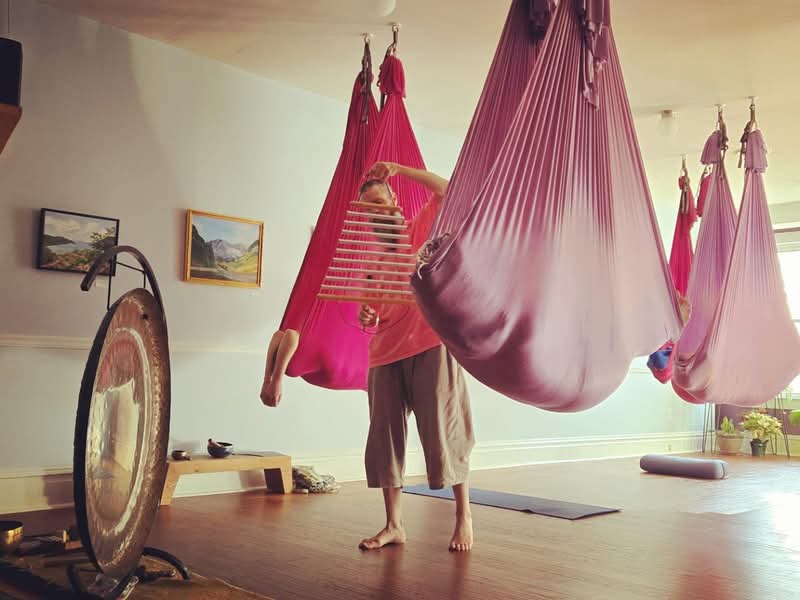 Person practicing aerial yoga in pink hammock, with other hammocks hanging in a bright room, wooden floor, and wall art.