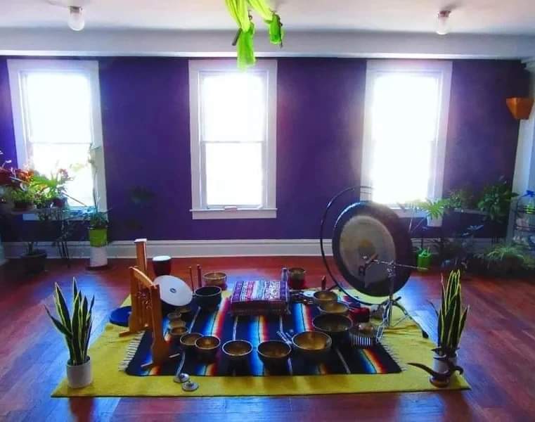 Indoor space with purple wall, three windows, plants, and a musical setup on a yellow mat, with a fan and chairs.