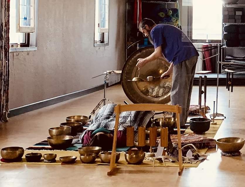 Person lying on the floor with bowls around, another person playing a gong nearby, in a room with windows and chairs.