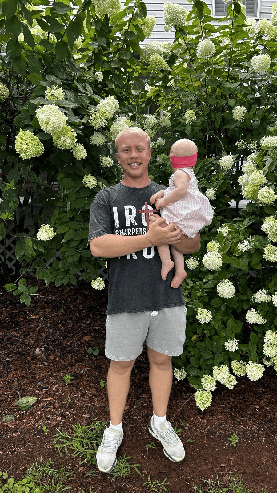 Young man holding a baby girl in front of white hydrangea flowers, smiling outdoors.