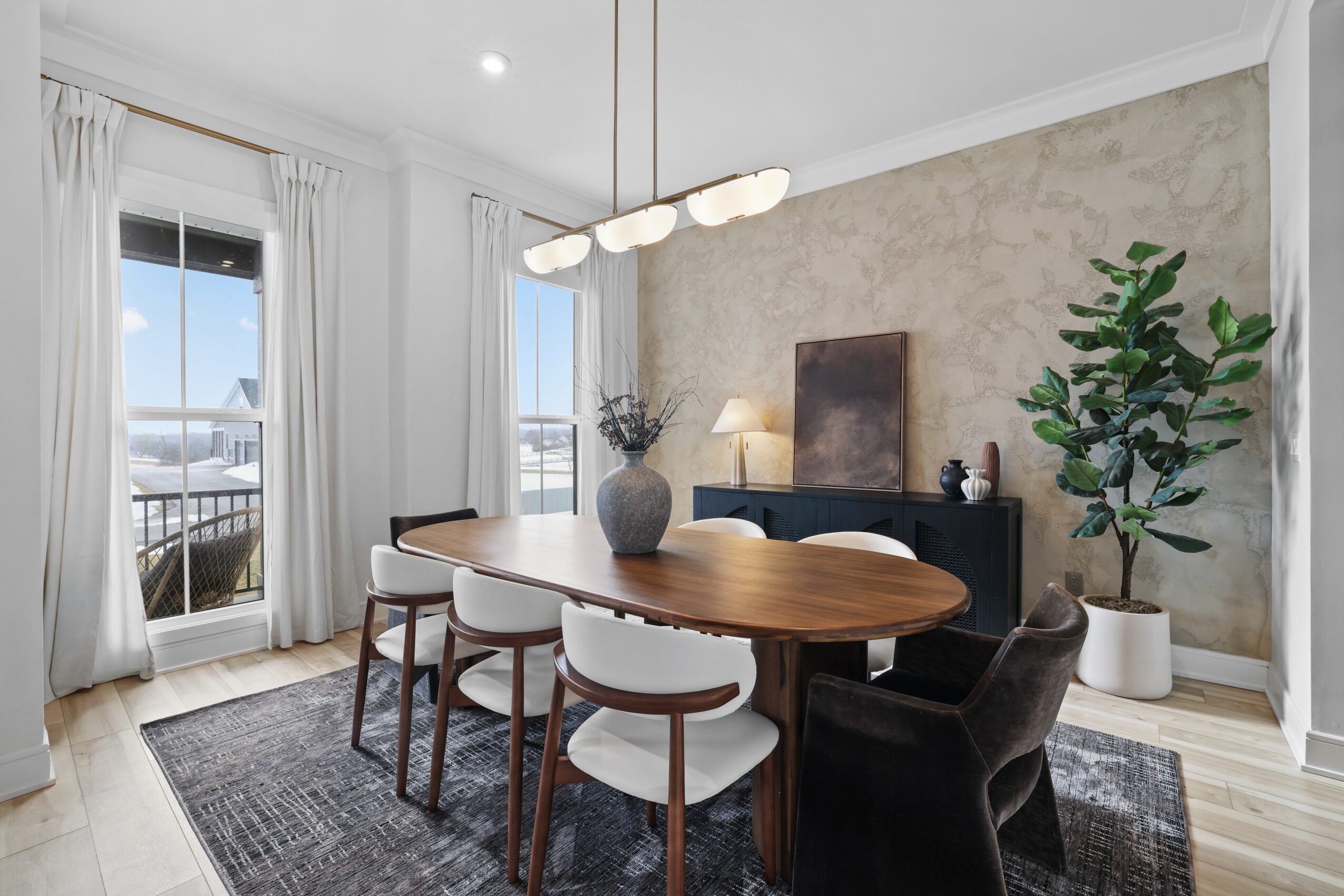 Dining room with oval wooden table, six chairs, a sideboard, large window, and potted plant, bright and modern decor.