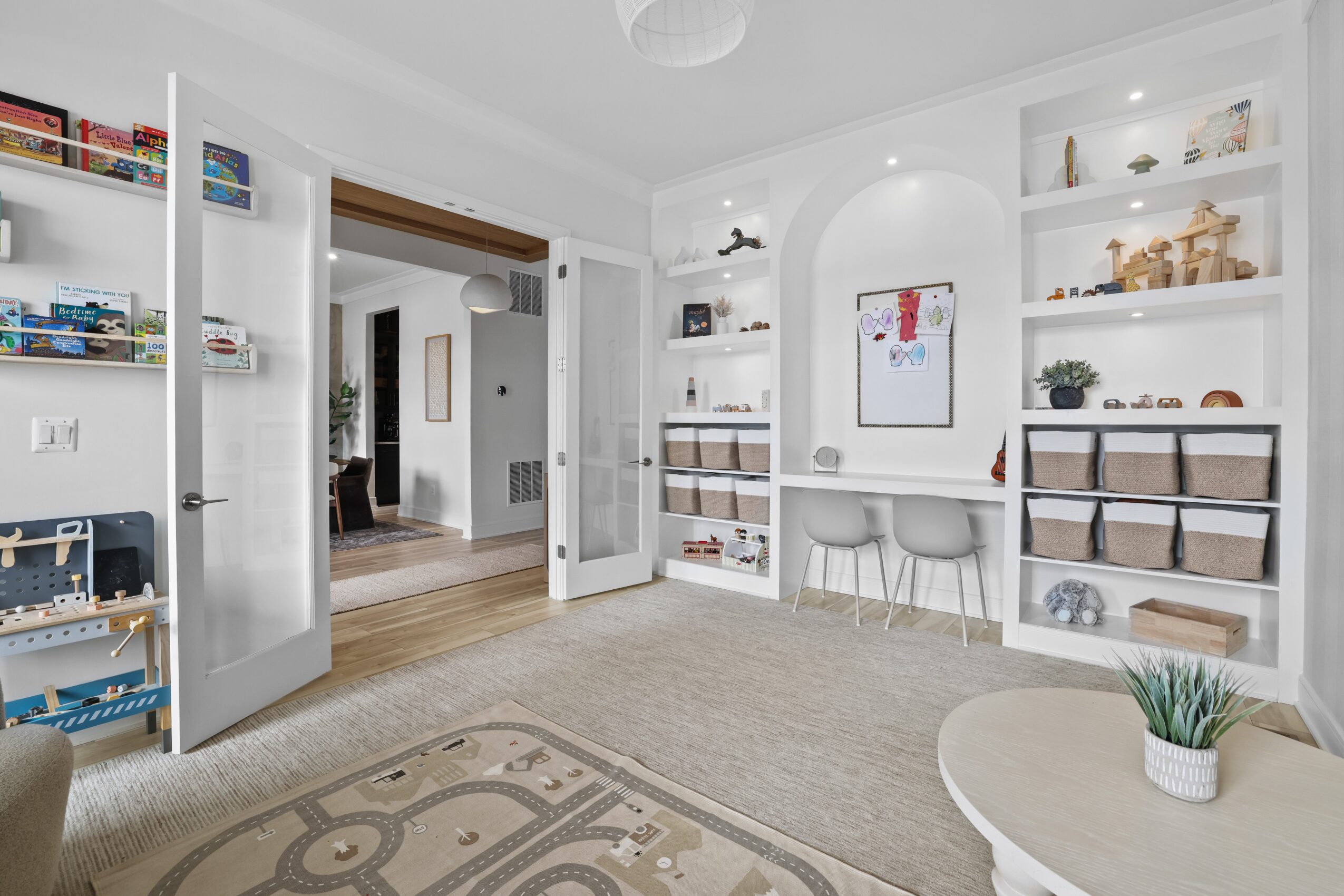 Living room with white built-in shelves, a beige carpet, and a round table with a plant, open door to another room