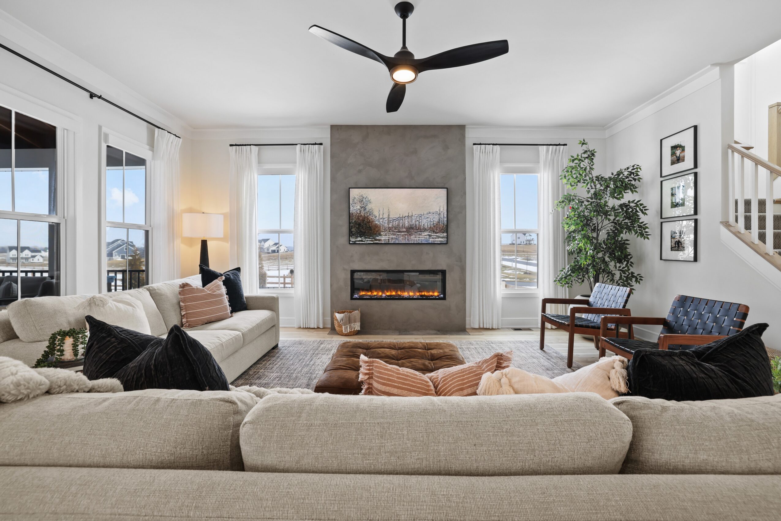 Living room with a large beige sofa, fireplace, wall-mounted TV, and large windows, decorated with plants and framed pictures.