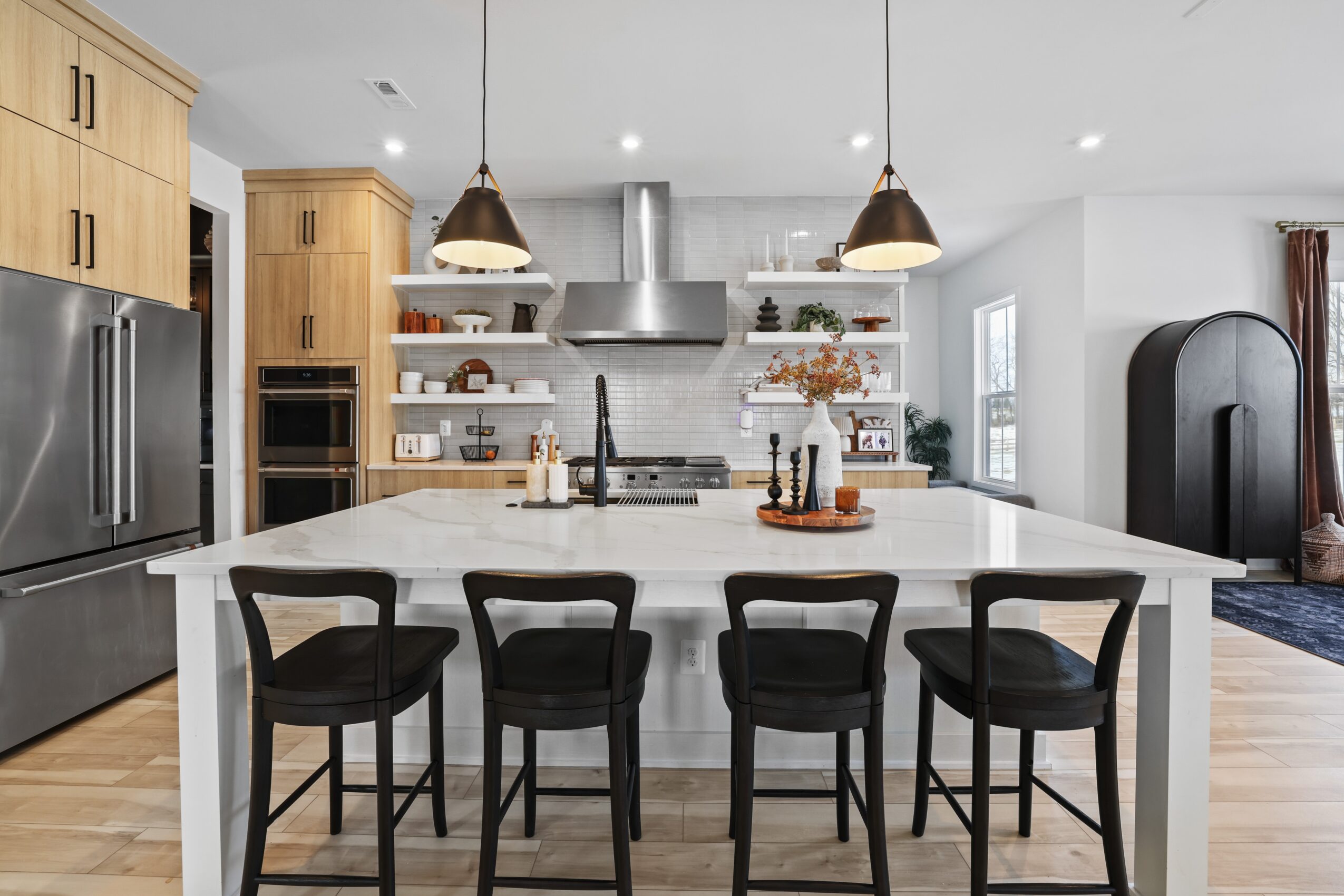Modern kitchen with white island, black chairs, wooden cabinets, stainless steel appliances, and pendant lights, bright and spacious.