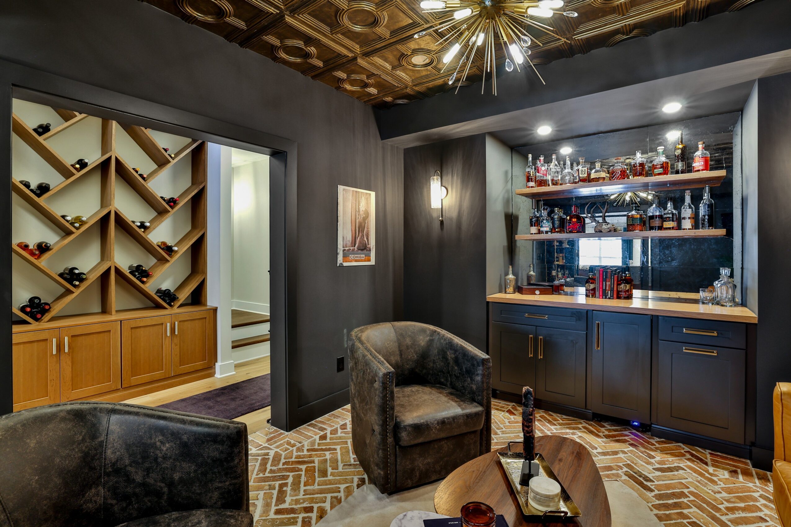Interior of a bar with dark walls, a shelf of bottles, two armchairs, and a wooden floor.