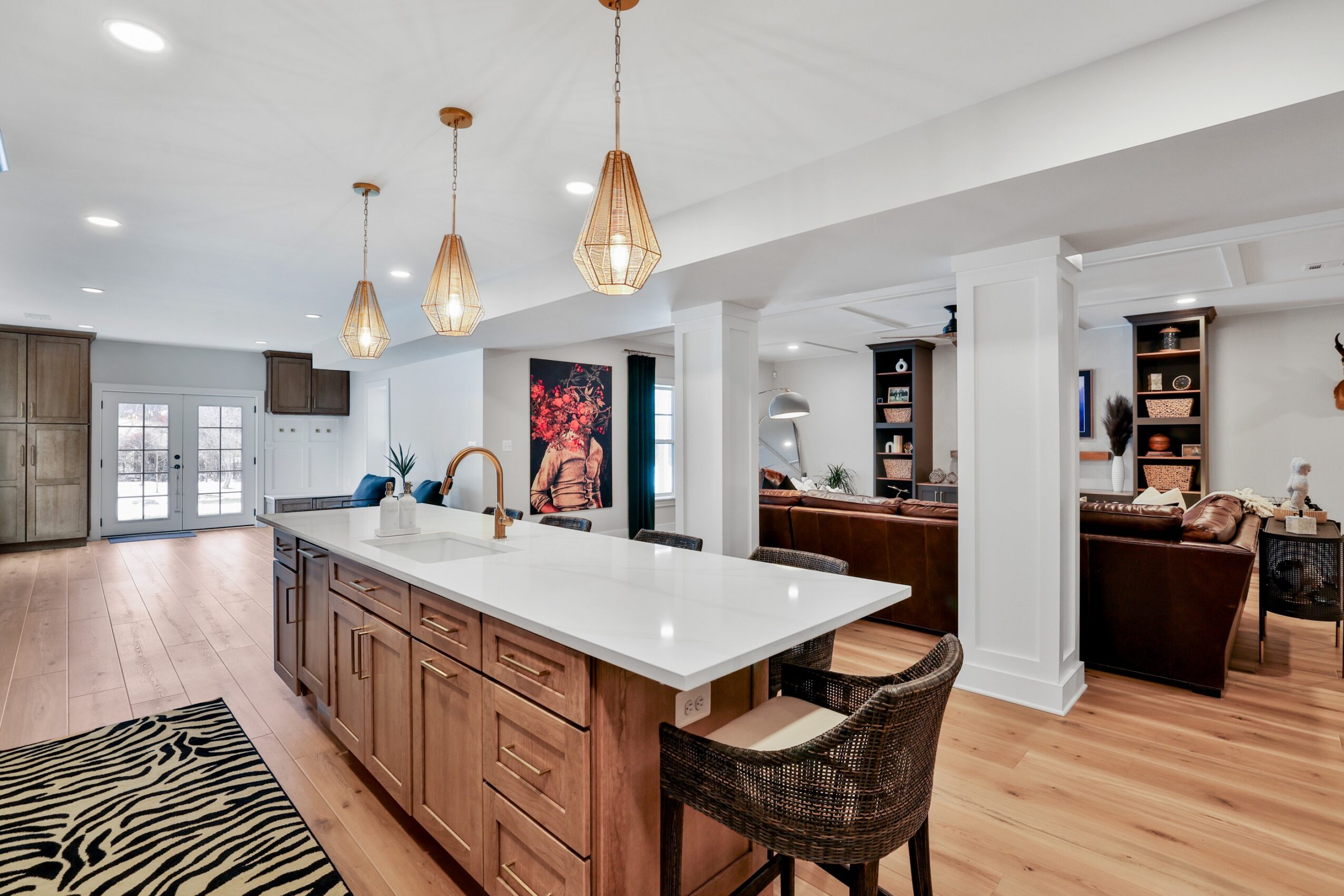 Open kitchen with white island, pendant lights, and living area with sofas and bookshelves, hardwood floors, and large windows.