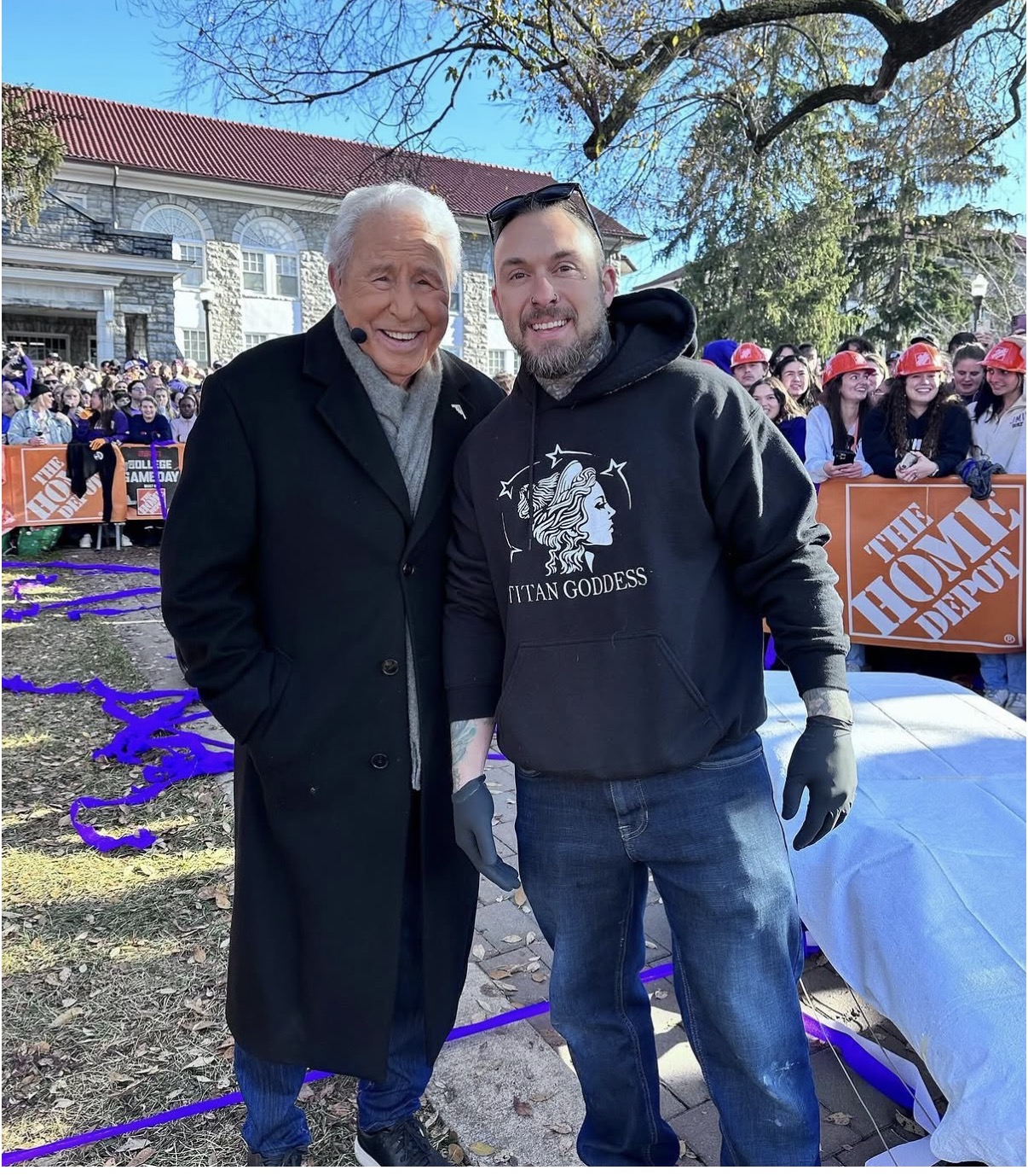 Two men smiling outdoors, one elderly with white hair in a dark coat, the other younger in a hoodie, with a crowd and trees in background.