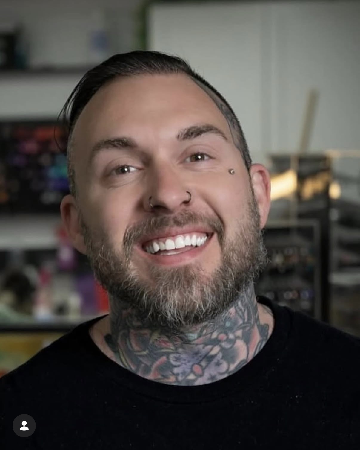 Smiling man with a beard and tattoos on neck, in a room with shelves and equipment in background.