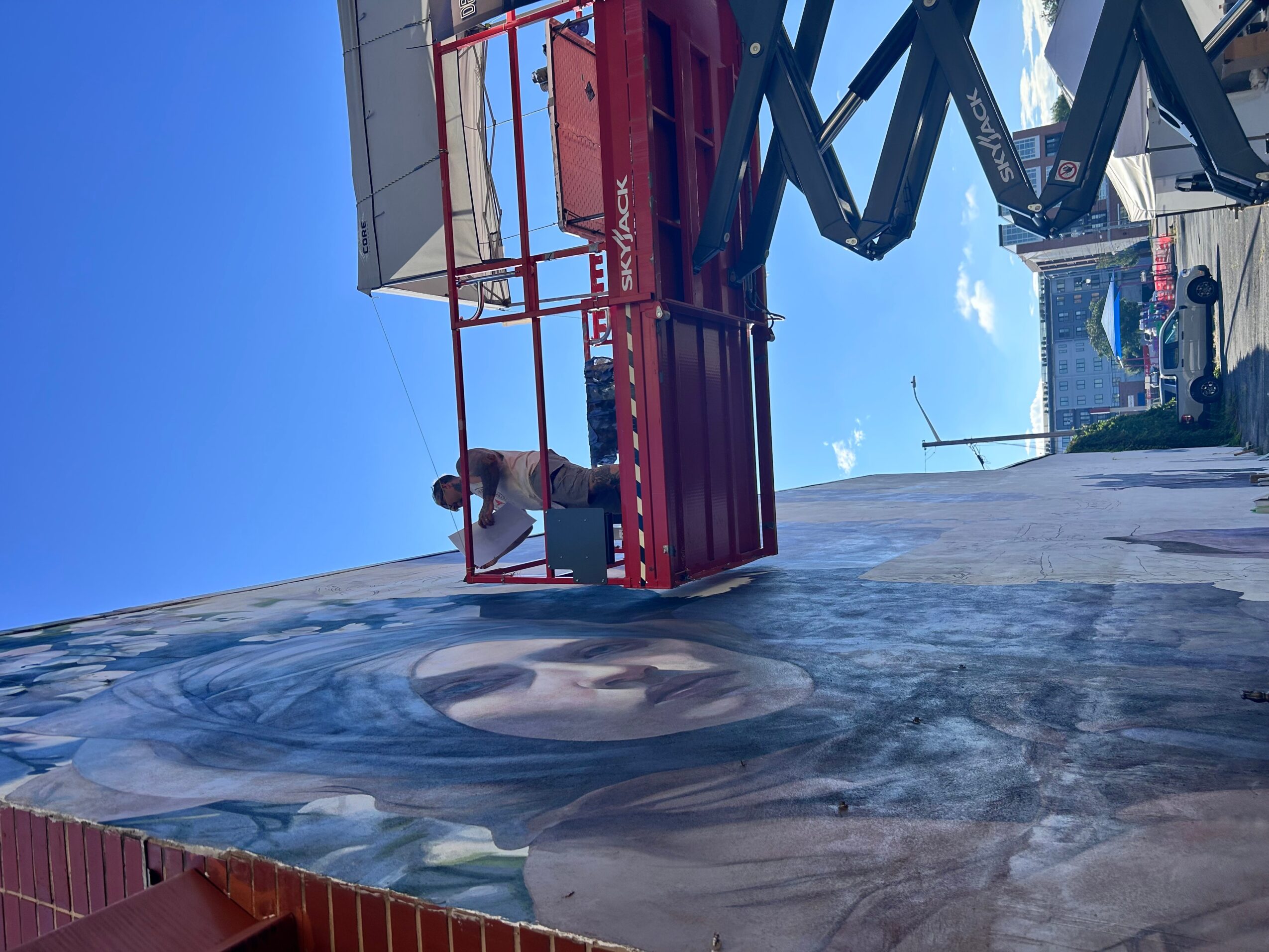 Person on a red scissor lift working on a mural wall, with a blue sky background.