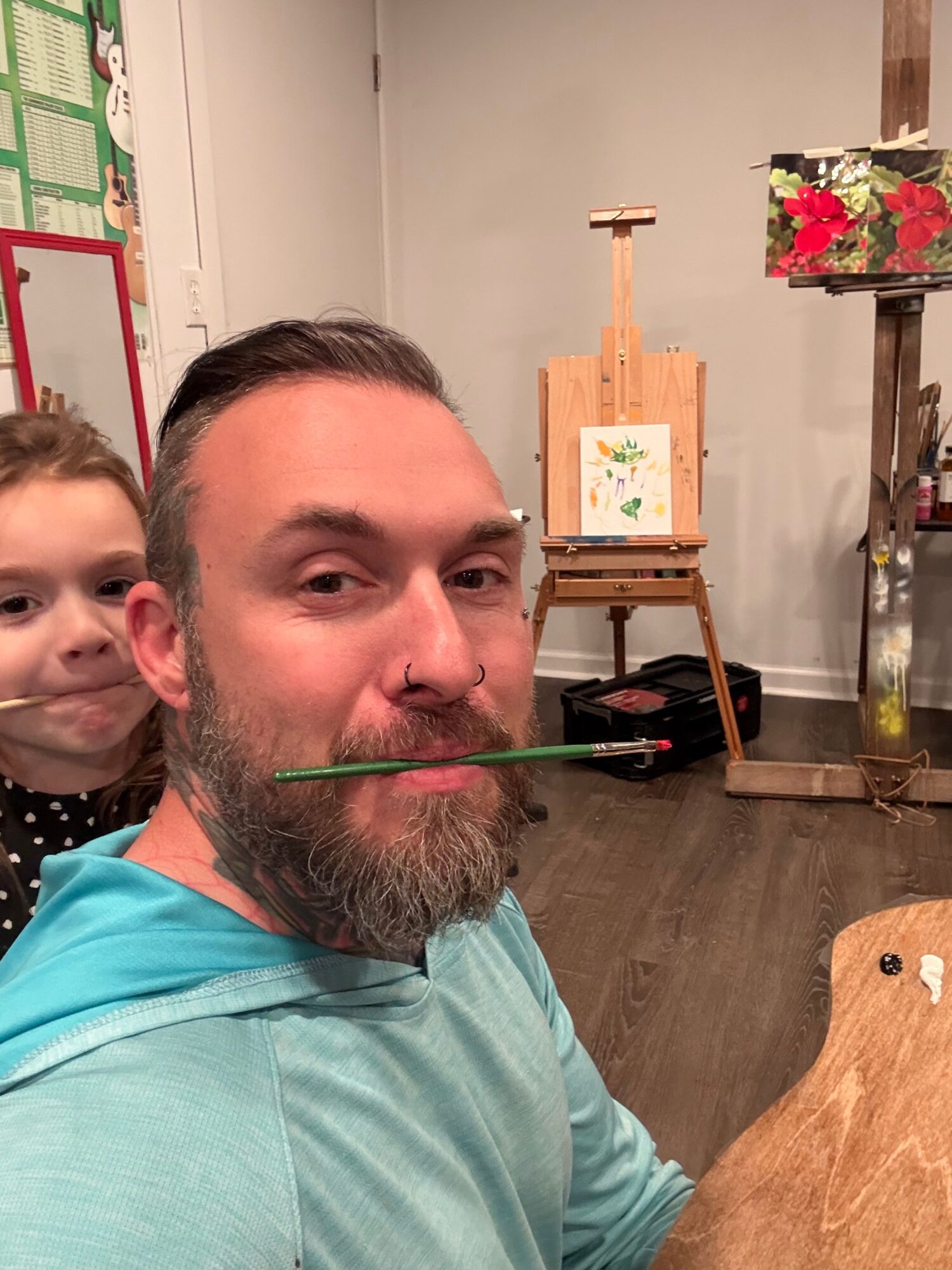 Man with a beard and a woman with curly hair sit indoors, with an easel and flowers in the background.