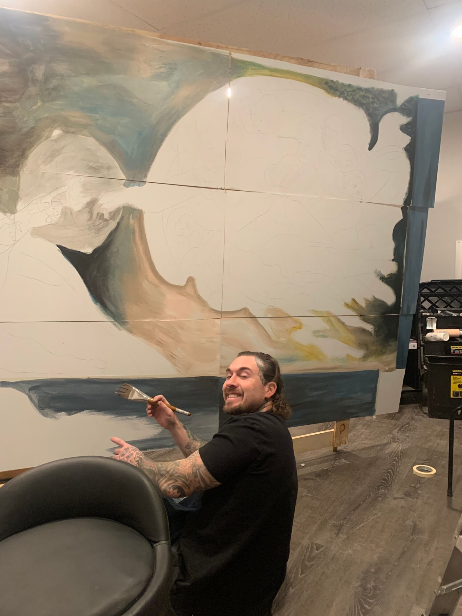 Person with dark hair and beard pointing at a large abstract mural on a wall, with a black chair nearby.