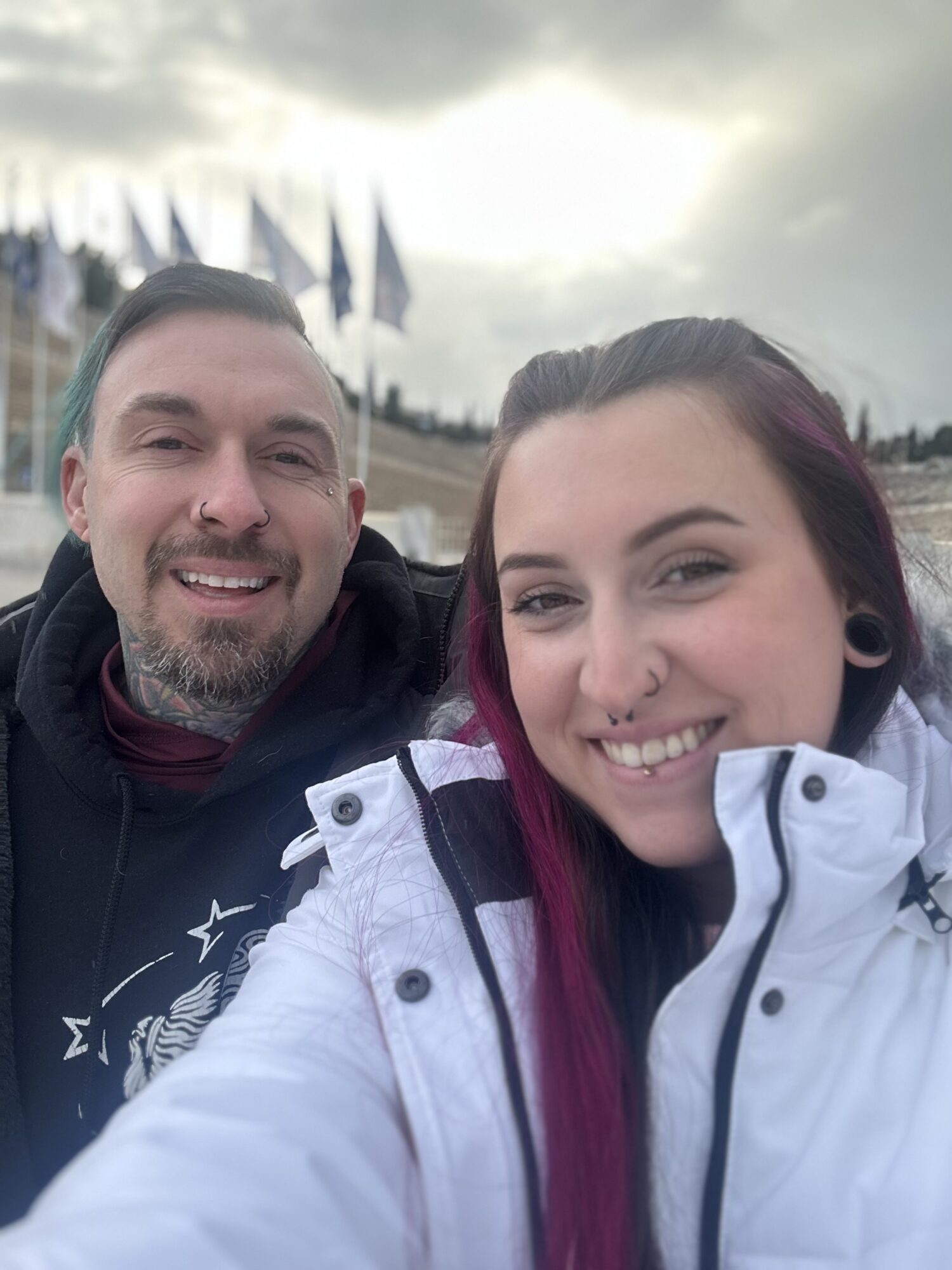 Two people smiling outdoors with flags and cloudy sky in background.