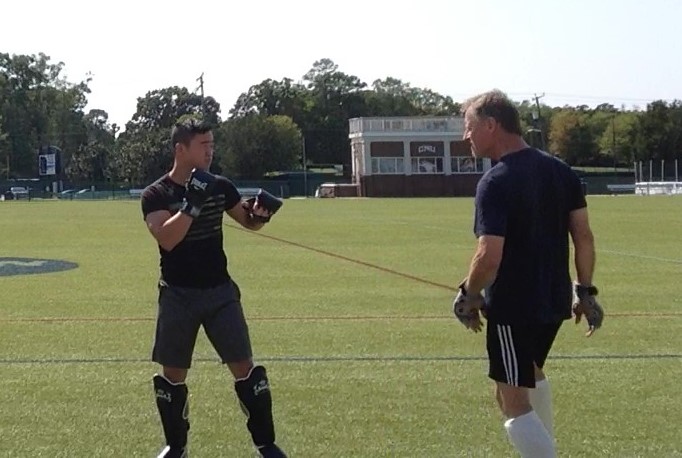Two people on a sports field, one holding a training tool, with trees and a building in the background.