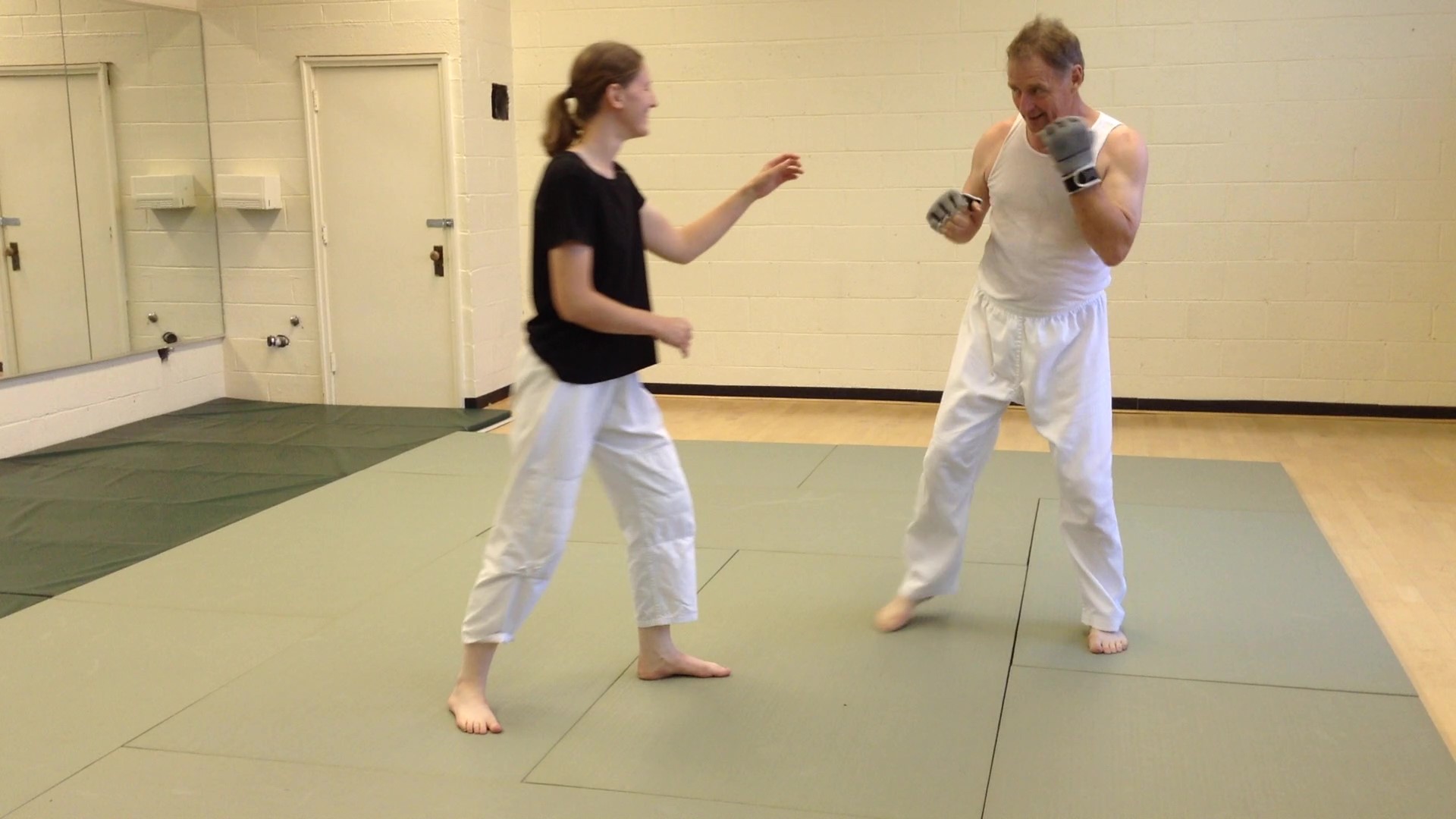 Two people practicing martial arts on a mat, one in a black shirt and white pants, the other in a white outfit, in a room with a mirror and door.