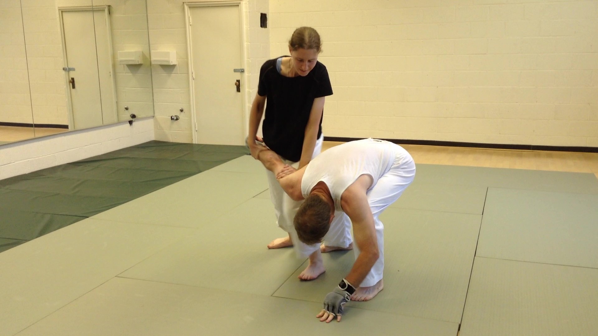 Two people practicing a grappling move on a mat in a room with white walls and a mirror, one in black, one in white.