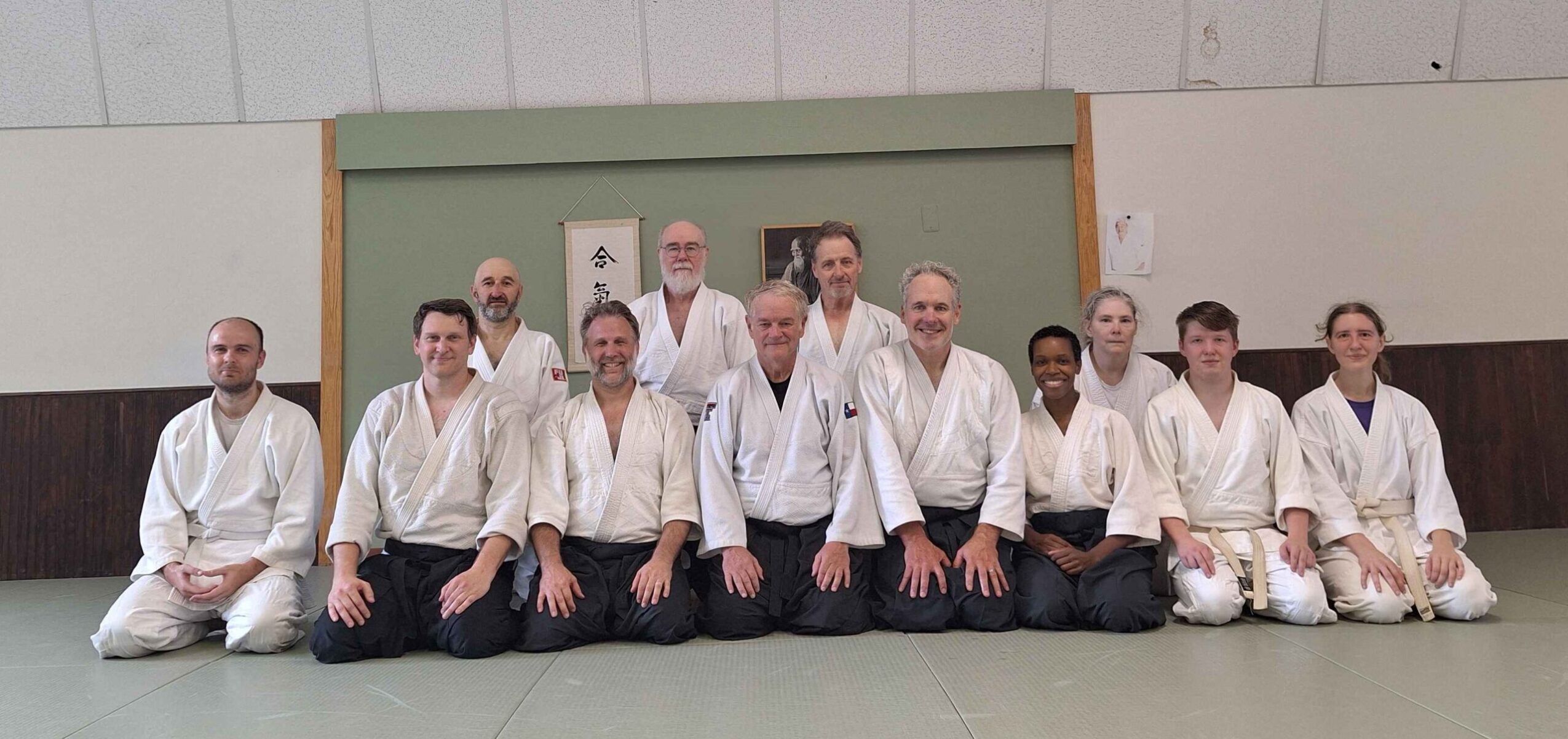 Group of people in martial arts uniforms kneeling on mat in dojo, with green and white wall background.