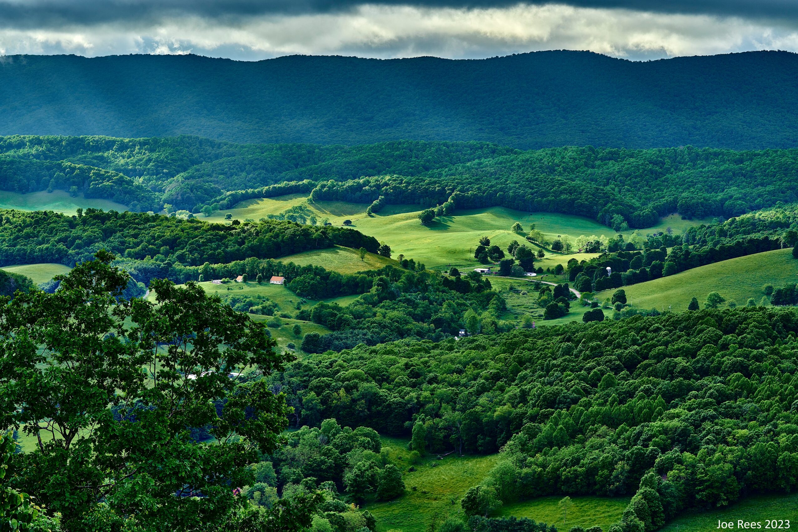 Lush green rolling hills with trees and fields under a partly cloudy sky, distant mountains in the background.