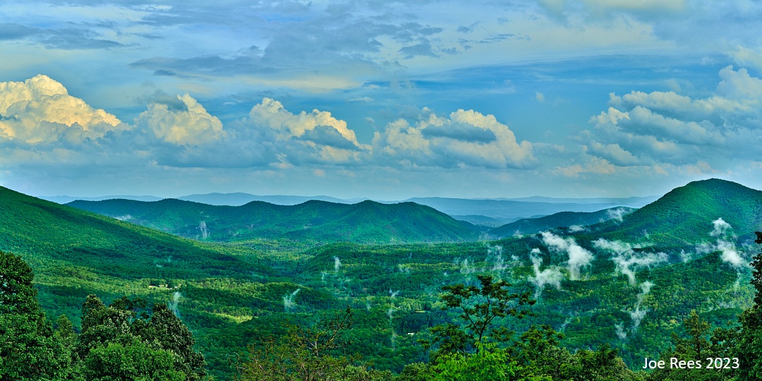 Scenic landscape with green hills, mountains, and a partly cloudy sky, with some smoke rising from the forest.