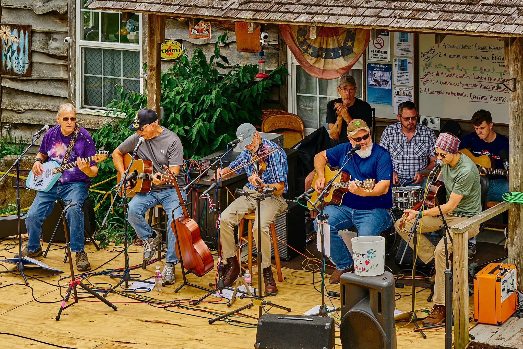 Group of eight musicians playing guitars and singing outdoors, with colorful decorations and a mural in the background.
