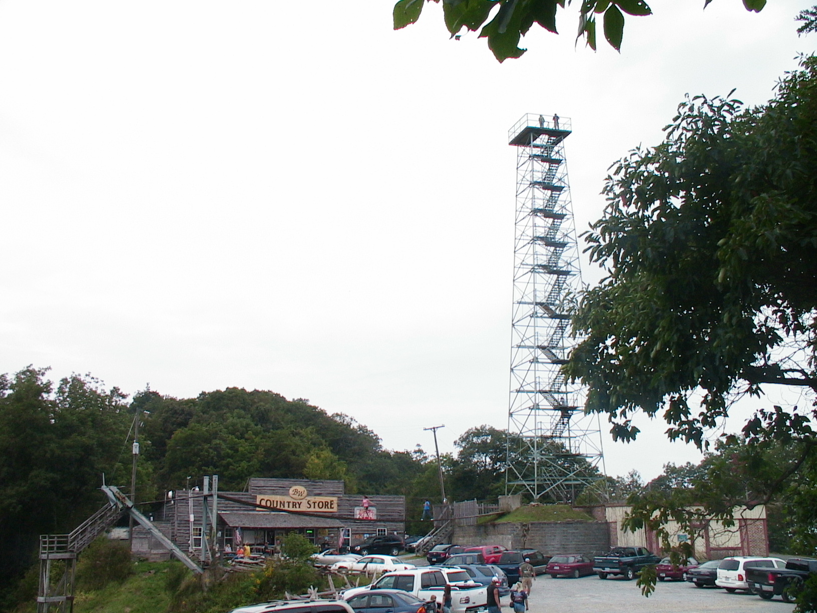 Parking lot with cars, trees, and a tall metal tower against a cloudy sky.