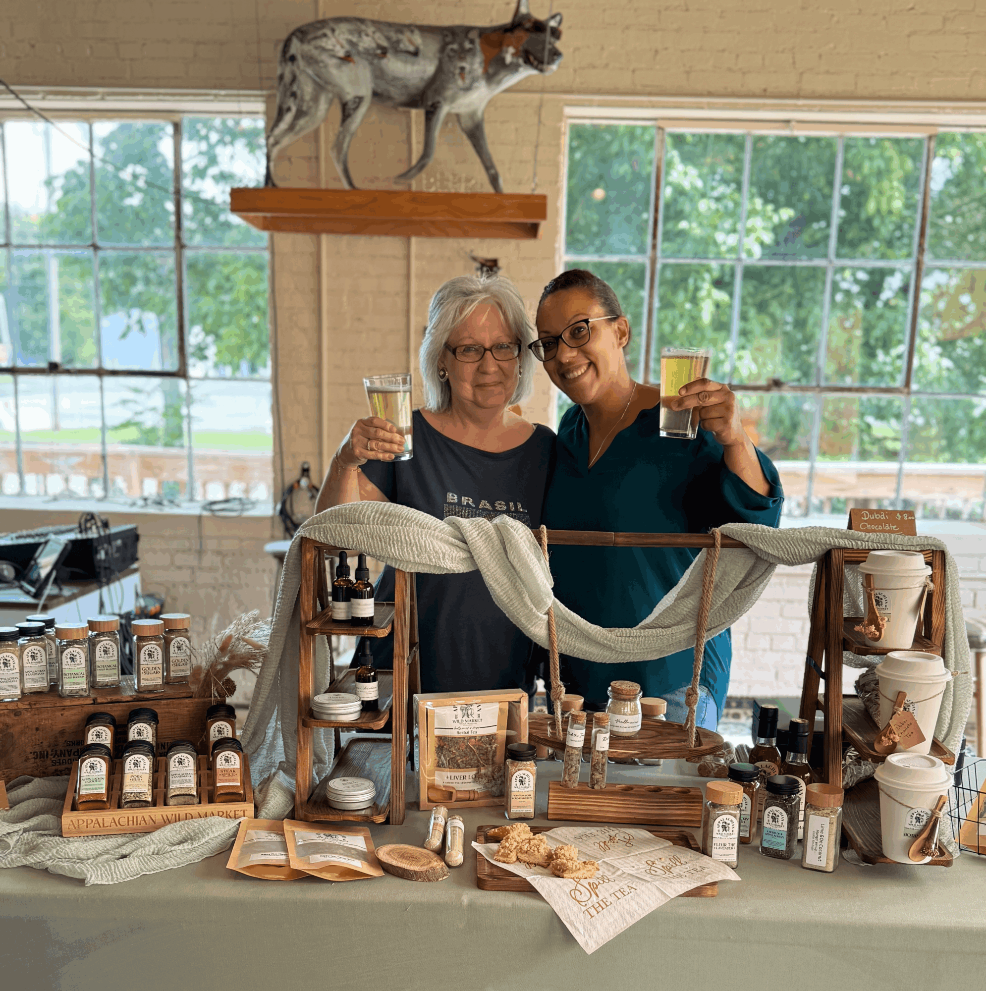 Two women stand behind a table with jars and products, holding glasses, inside a bright room with large windows and a mounted animal on the wall.