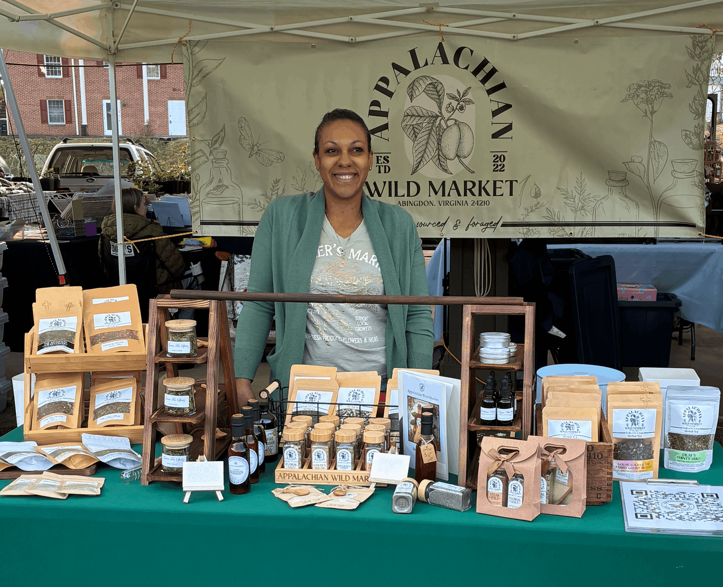 Smiling woman standing behind a table with jars and products at an outdoor market stall, with a banner in the background.