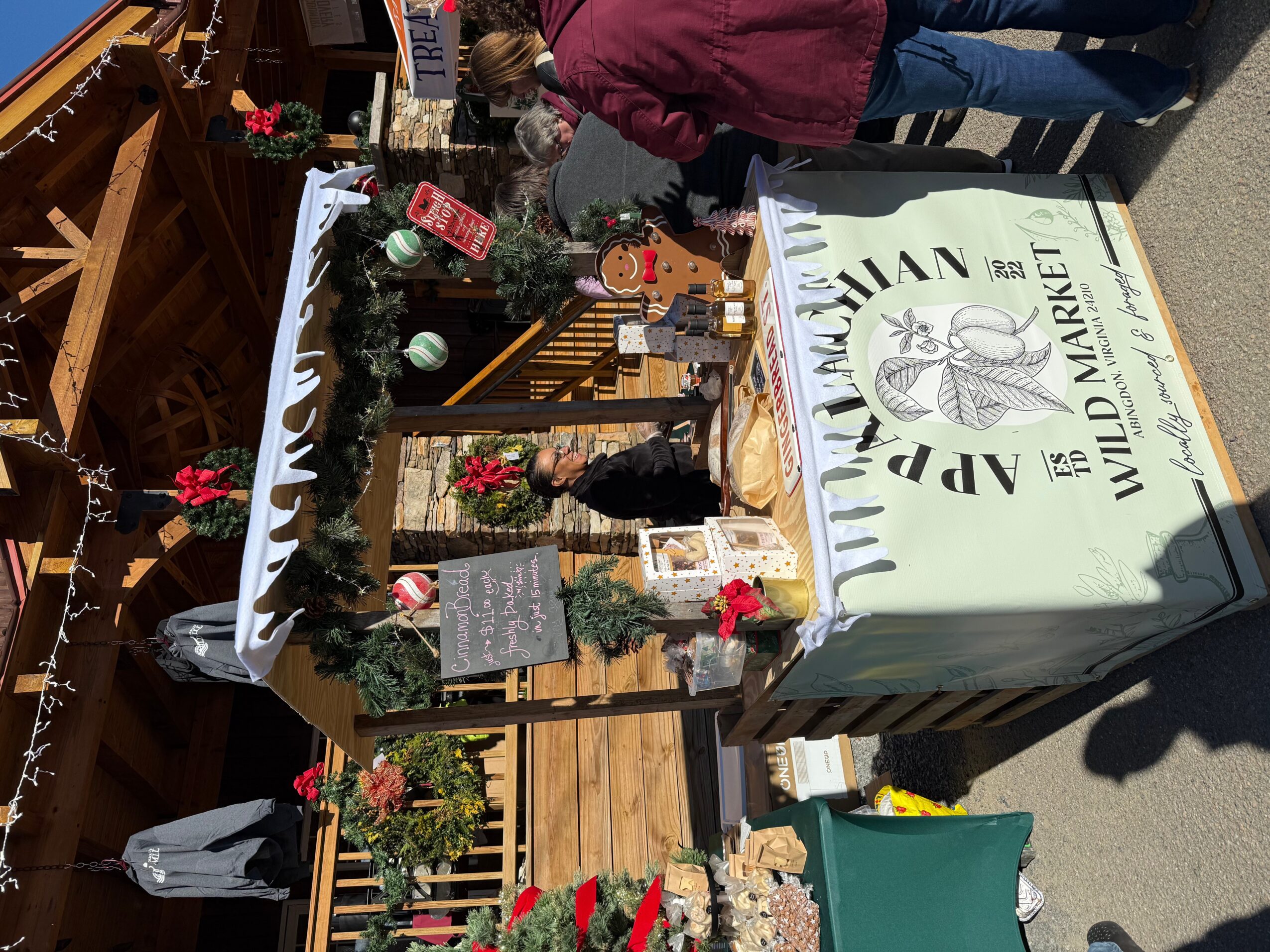 Table with holiday decorations, small trees, and a sign for a market, outdoors with people nearby.