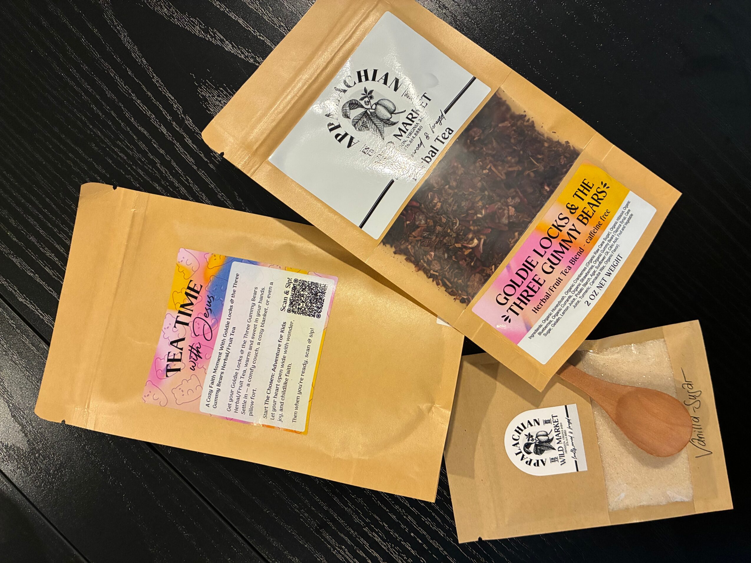 Three packages of tea and a wooden tea scoop on a black surface. One package has a transparent window showing tea leaves.