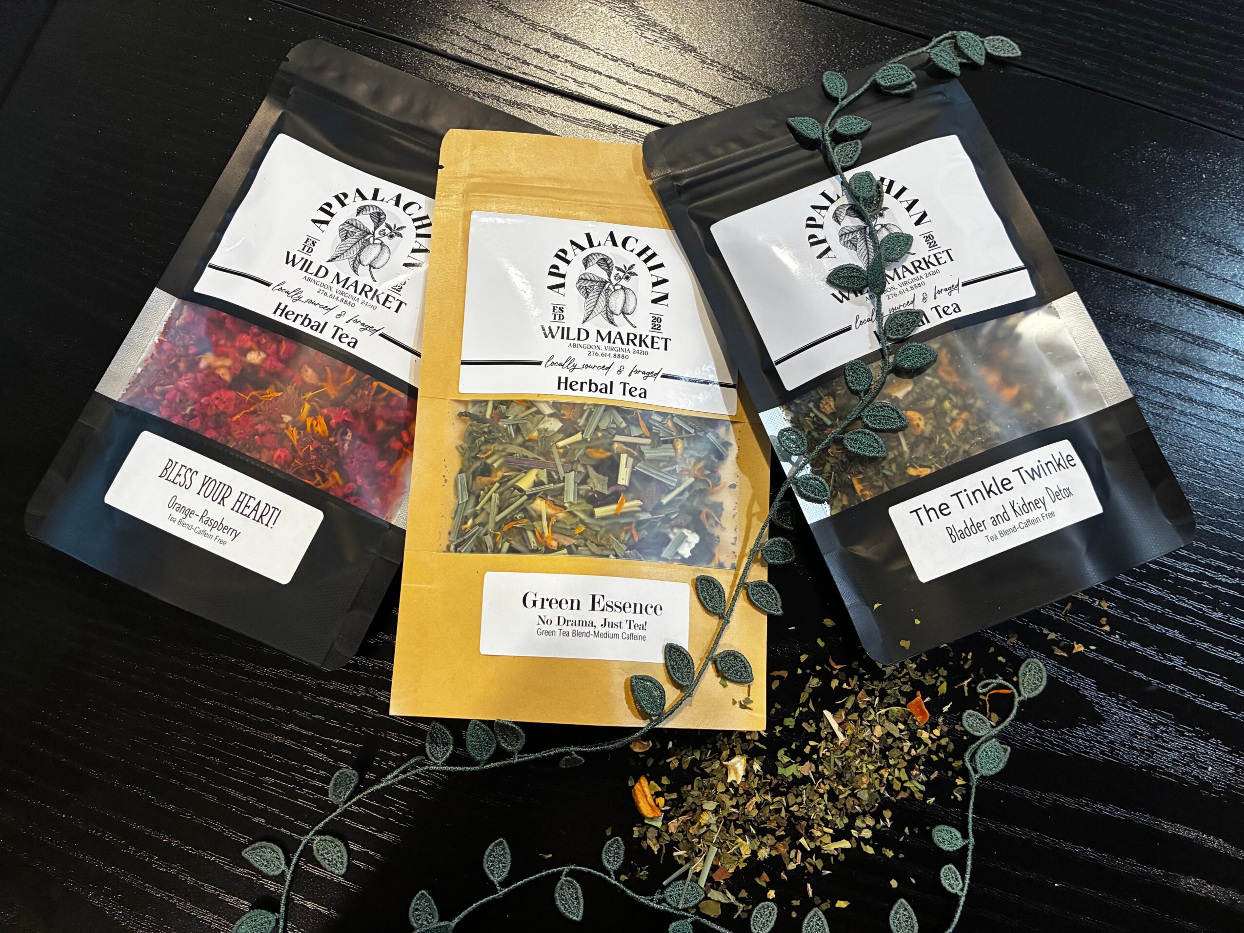 Three herbal tea packages with labels, arranged on a dark wooden surface, with loose herbs and a sprig of eucalyptus nearby.