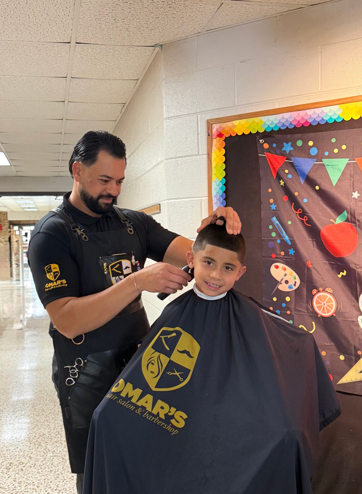 Man cutting a young boy's hair in a salon, boy wearing a black cape with yellow logo, colorful bulletin board in background.