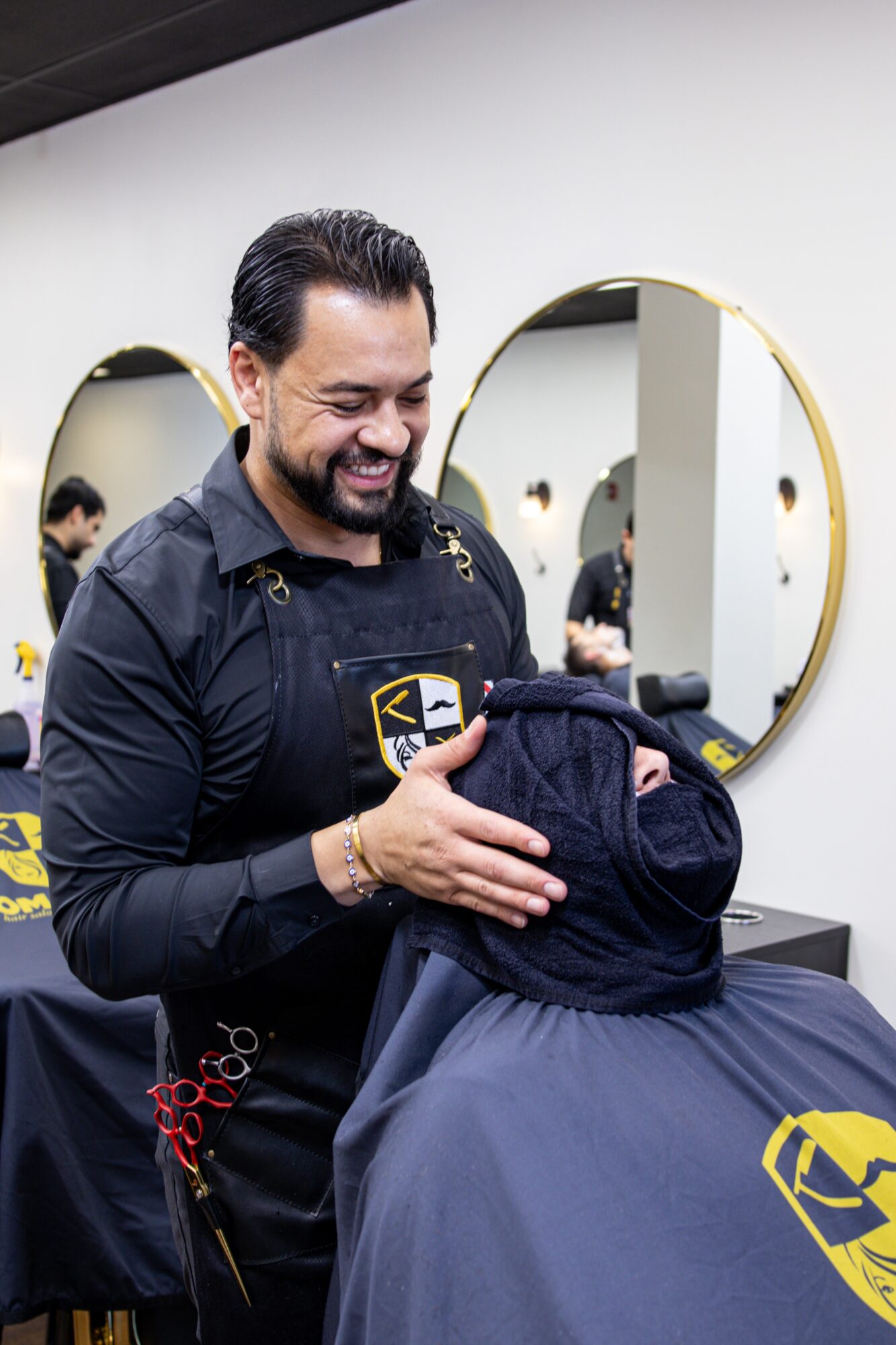 Man smiling and holding a backpack in a barbershop with mirrors and other people in the background.
