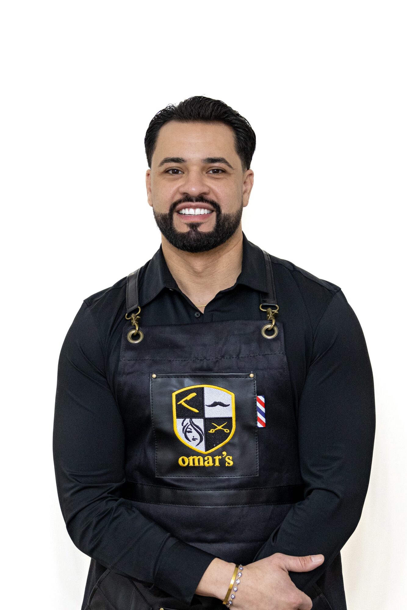 Man with dark hair and beard wearing a black shirt and apron, smiling, against a white background.