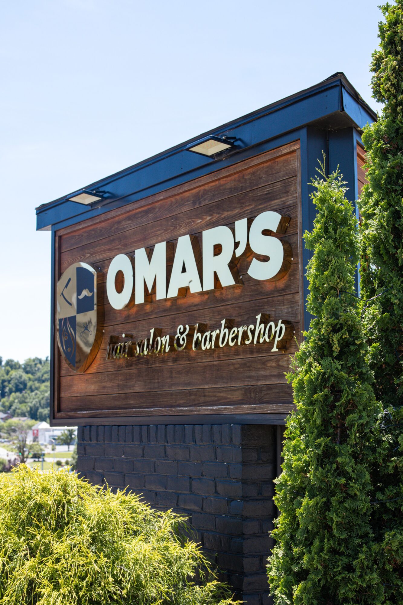 Sign for Omar's barbershop with a wooden background and white text, partially obscured by green trees.