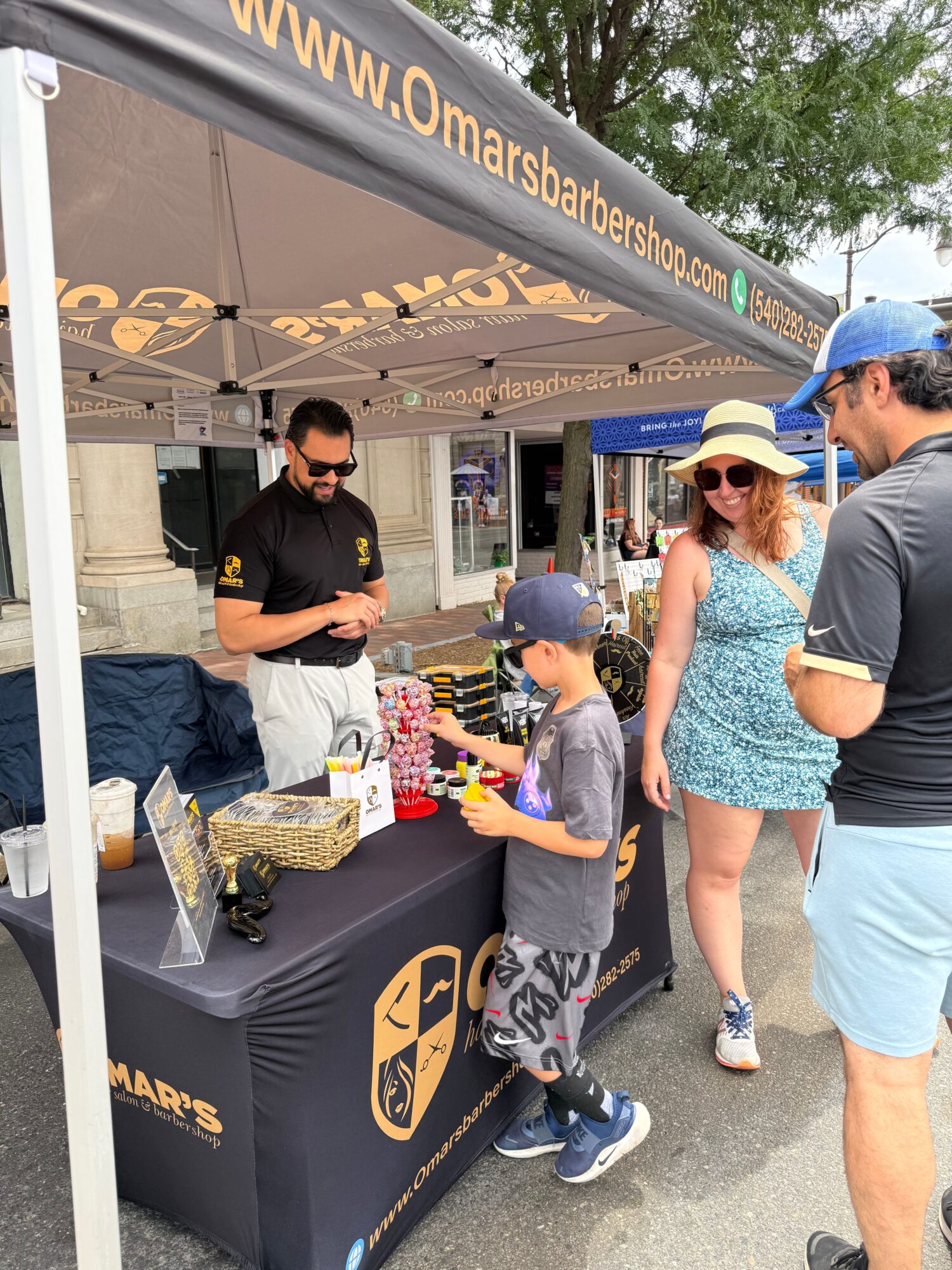 People gather around a table at an outdoor event, with a person in a black shirt and a person in a gray shirt engaging with items on the table.
