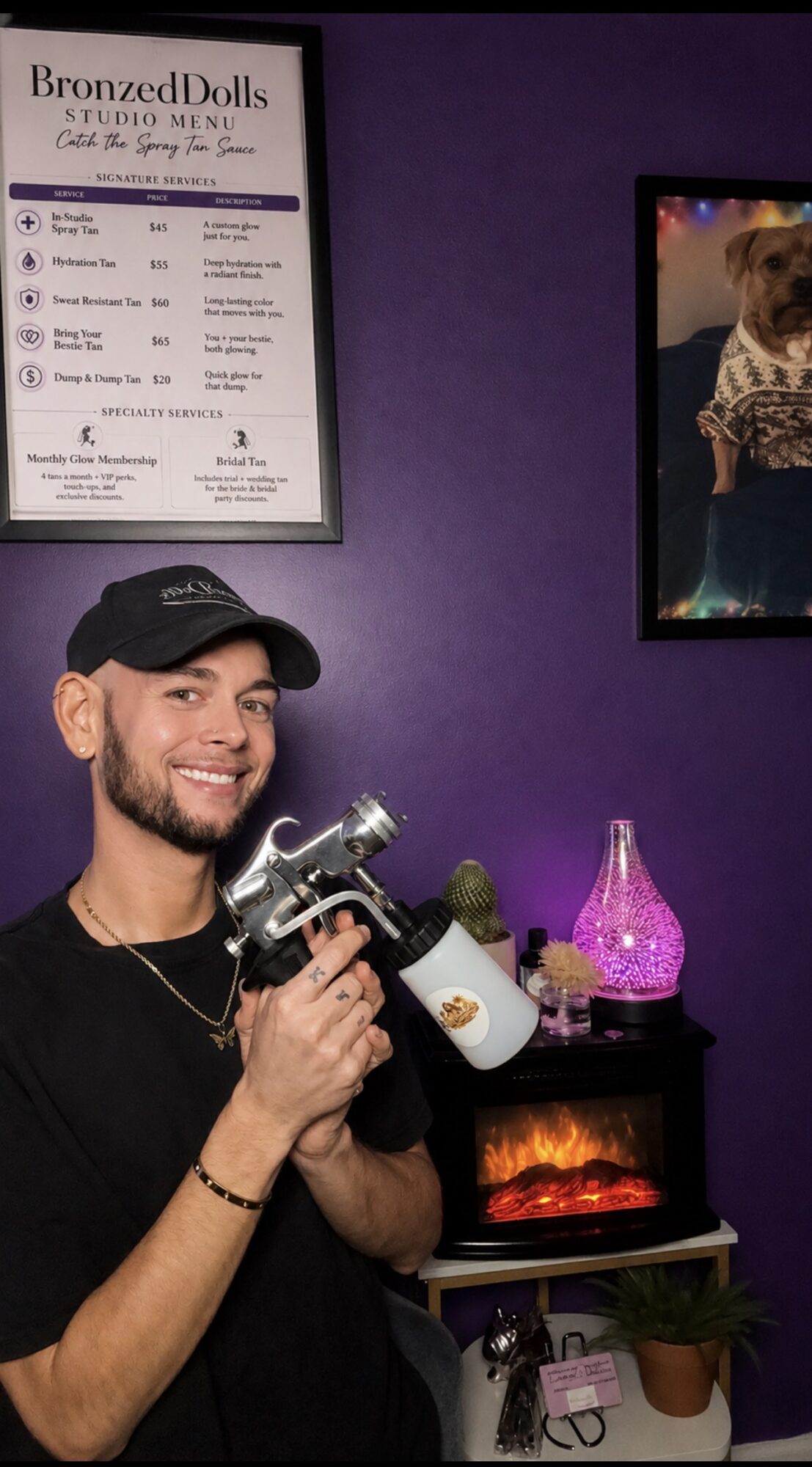 Smiling man wearing a black cap and shirt holding a tattoo machine in a room with purple walls, framed poster, and a pink lamp.