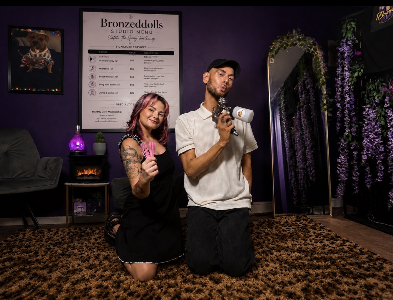Two people kneeling on a patterned carpet in a room with purple walls, a mirror, and decorative flowers, taking a selfie.