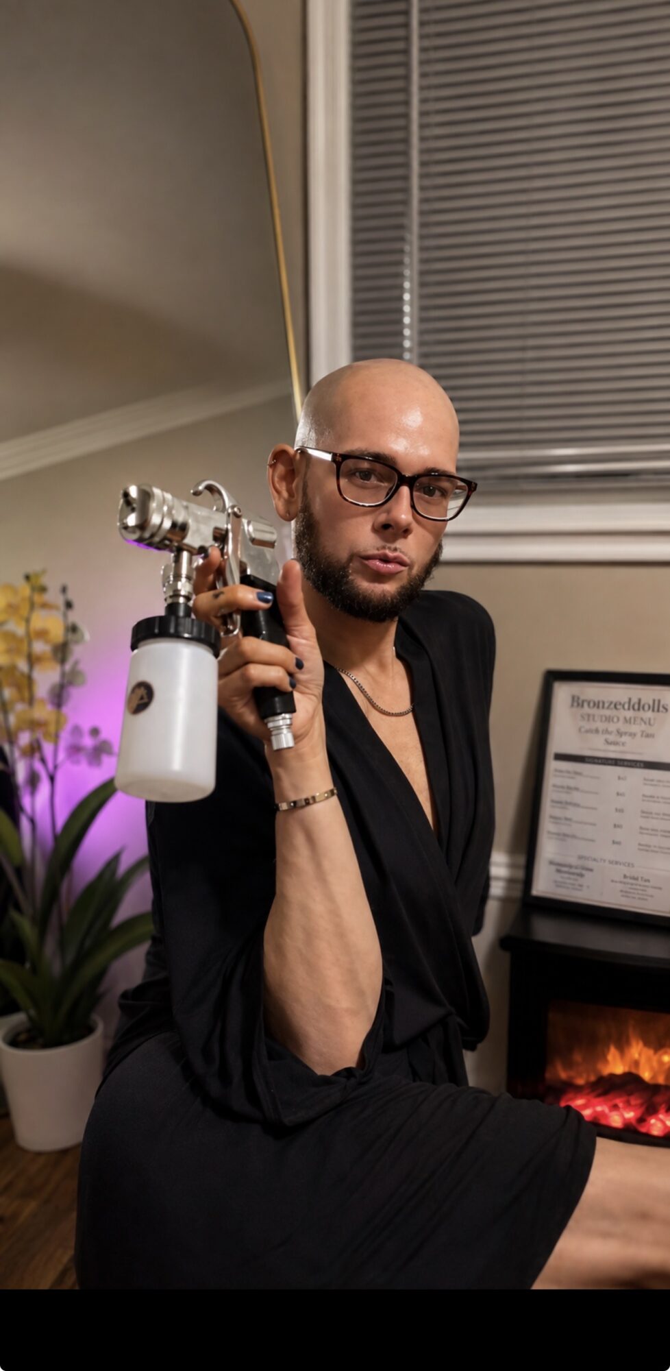 Person with glasses holding a spray gun, sitting indoors near a window and a potted plant.