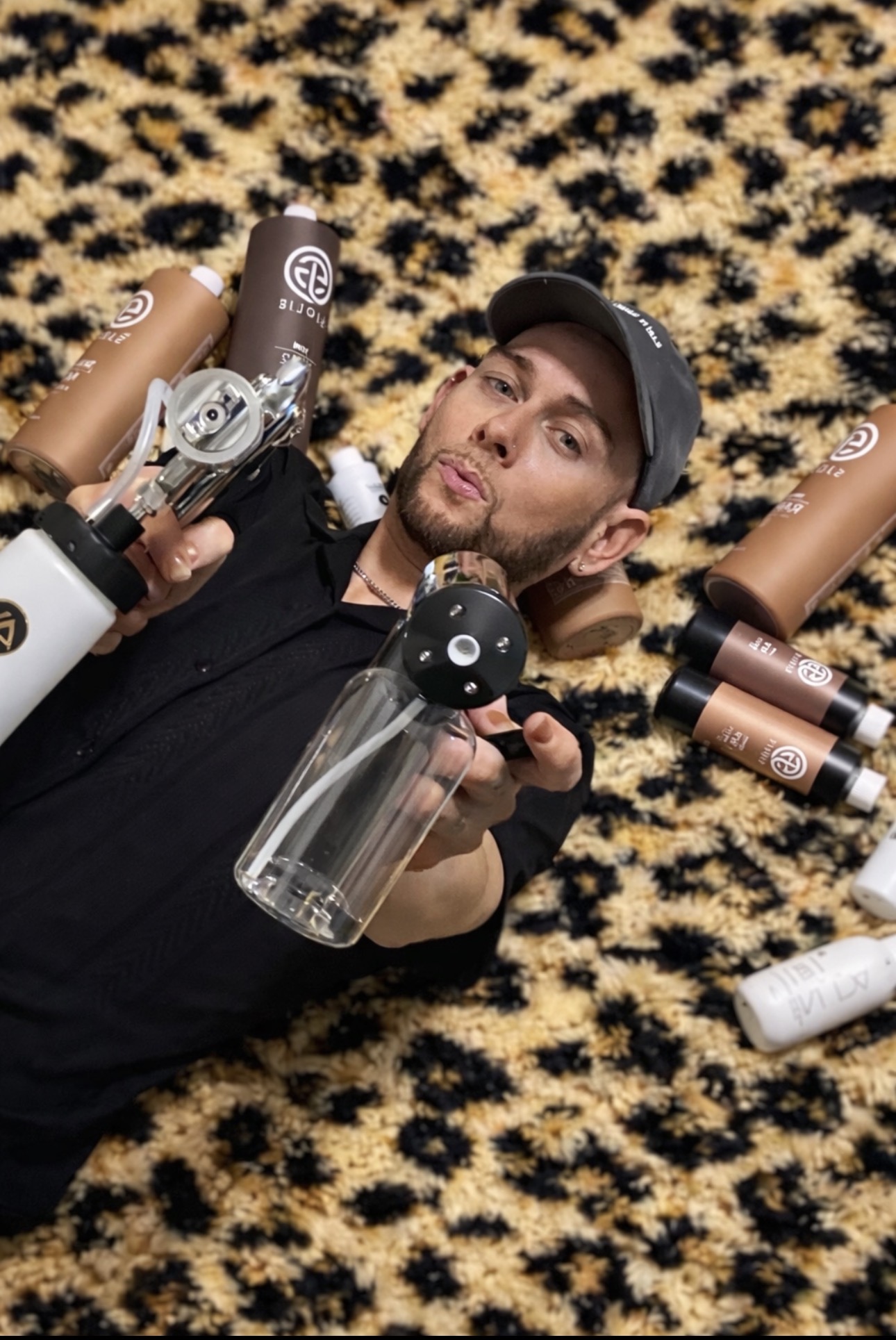 Person lying on a leopard print surface surrounded by spray bottles, holding a spray bottle toward the camera.