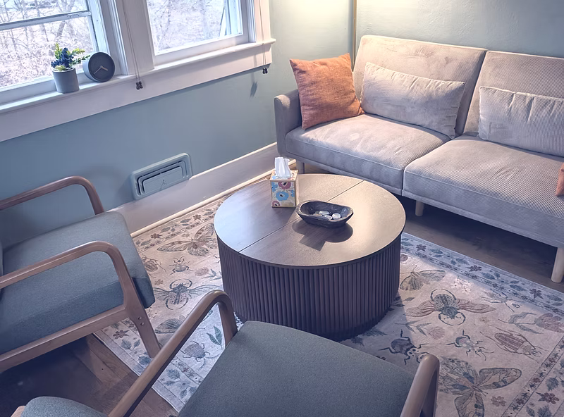Living room with a beige sofa, a round coffee table with a bottle and bowl, two chairs, a window with plants, and a patterned rug.