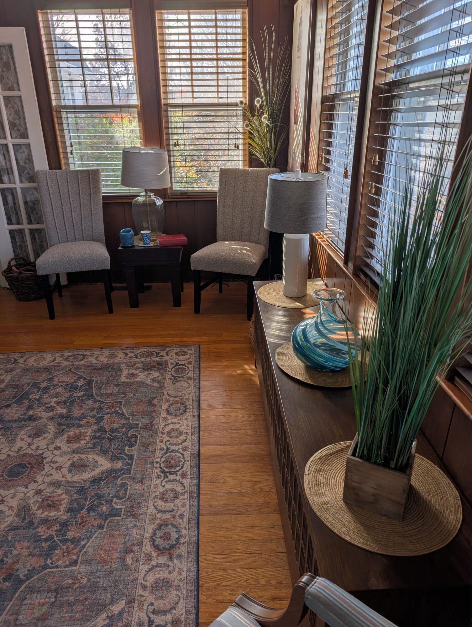 Sunlit room with two chairs, side table, lamps, and decorative vases near large windows with blinds.