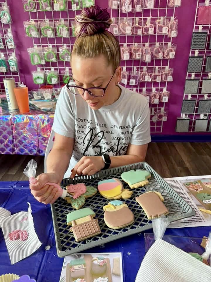 Young woman with glasses and a top knot decorating cookies on a table, with a pink wall and display in background.