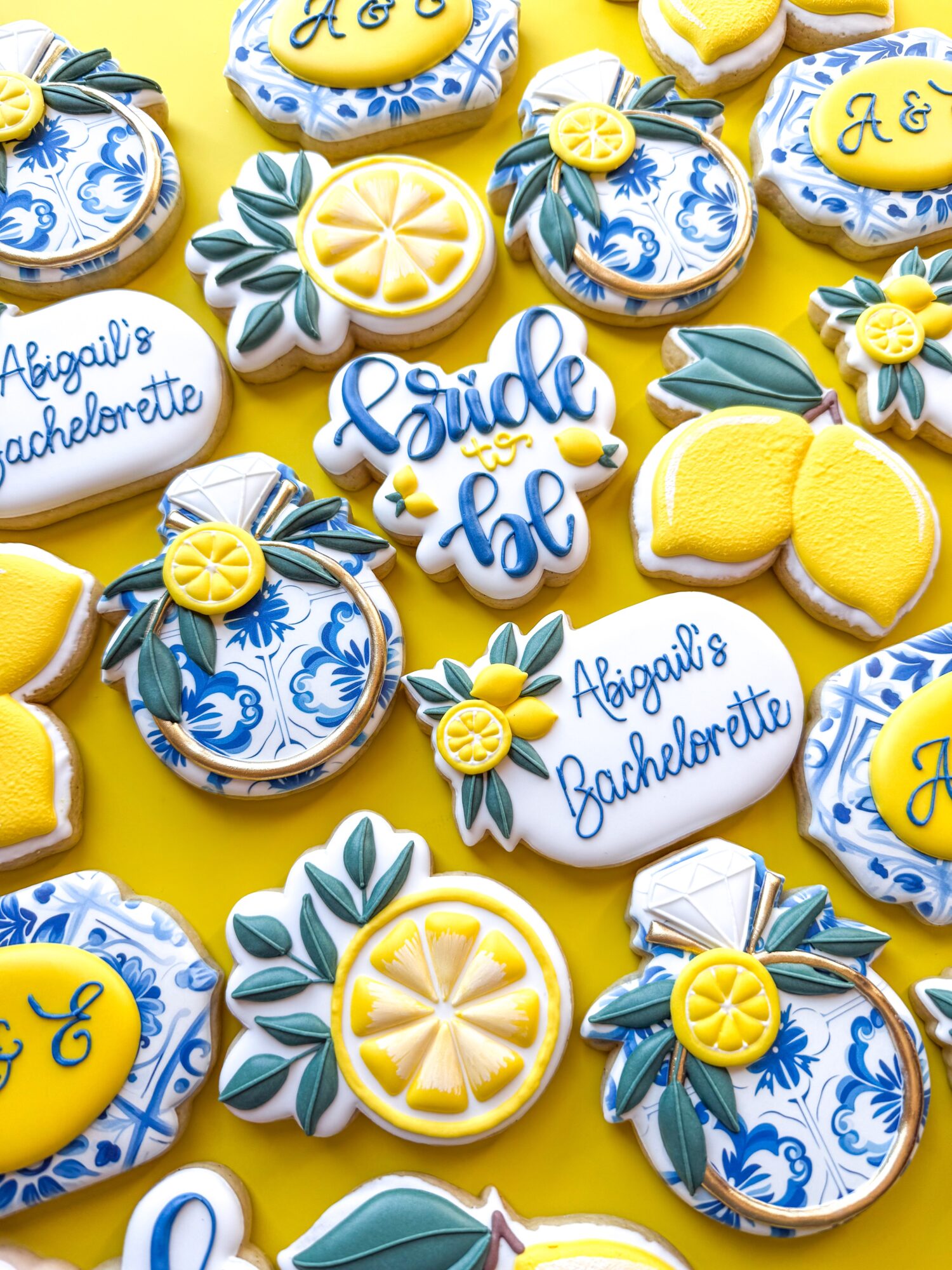 Decorative cookies with yellow, blue, and white designs, including lemons, flowers, and text for a bachelorette party, on a yellow background.