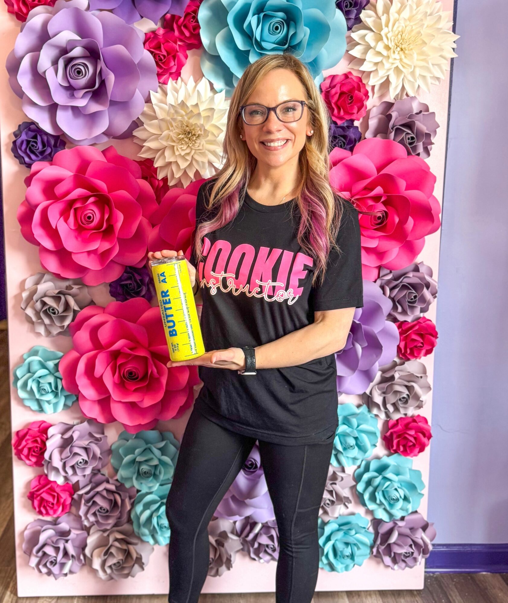 Woman with glasses holding a yellow can, standing in front of a colorful paper flower backdrop, smiling.